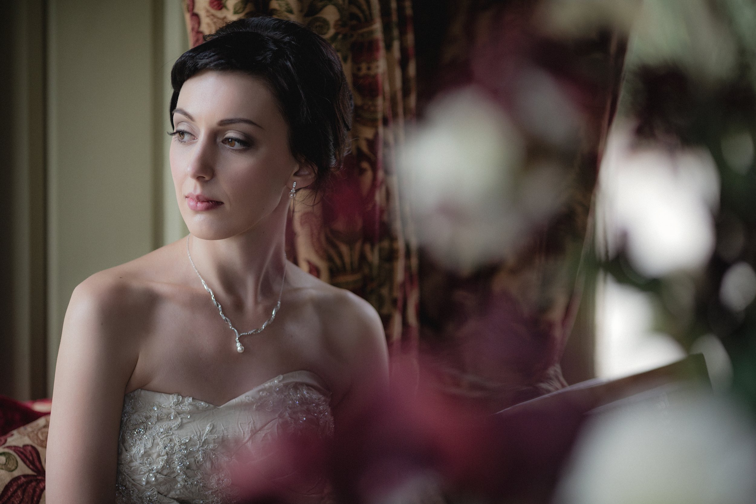 A young woman with short dark hair, wearing a strapless wedding dress with lace and beading, and a necklace with a pearl pendant, looks thoughtfully to the side inside a room with patterned curtains and floral decor.