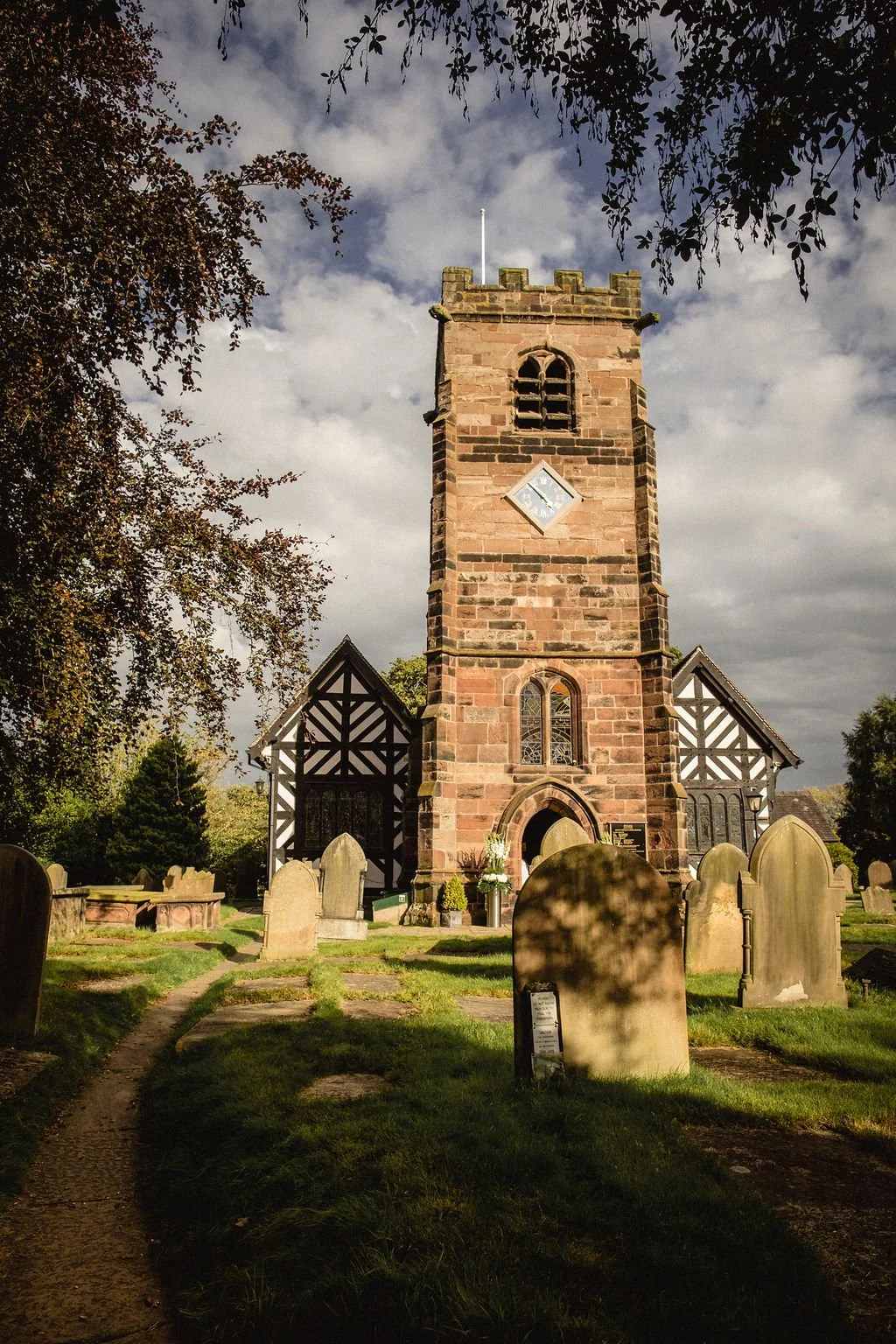A historic church with a tall stone tower, black and white timber framing on the sides, surrounded by a graveyard with headstones, green trees, and a cloudy sky.