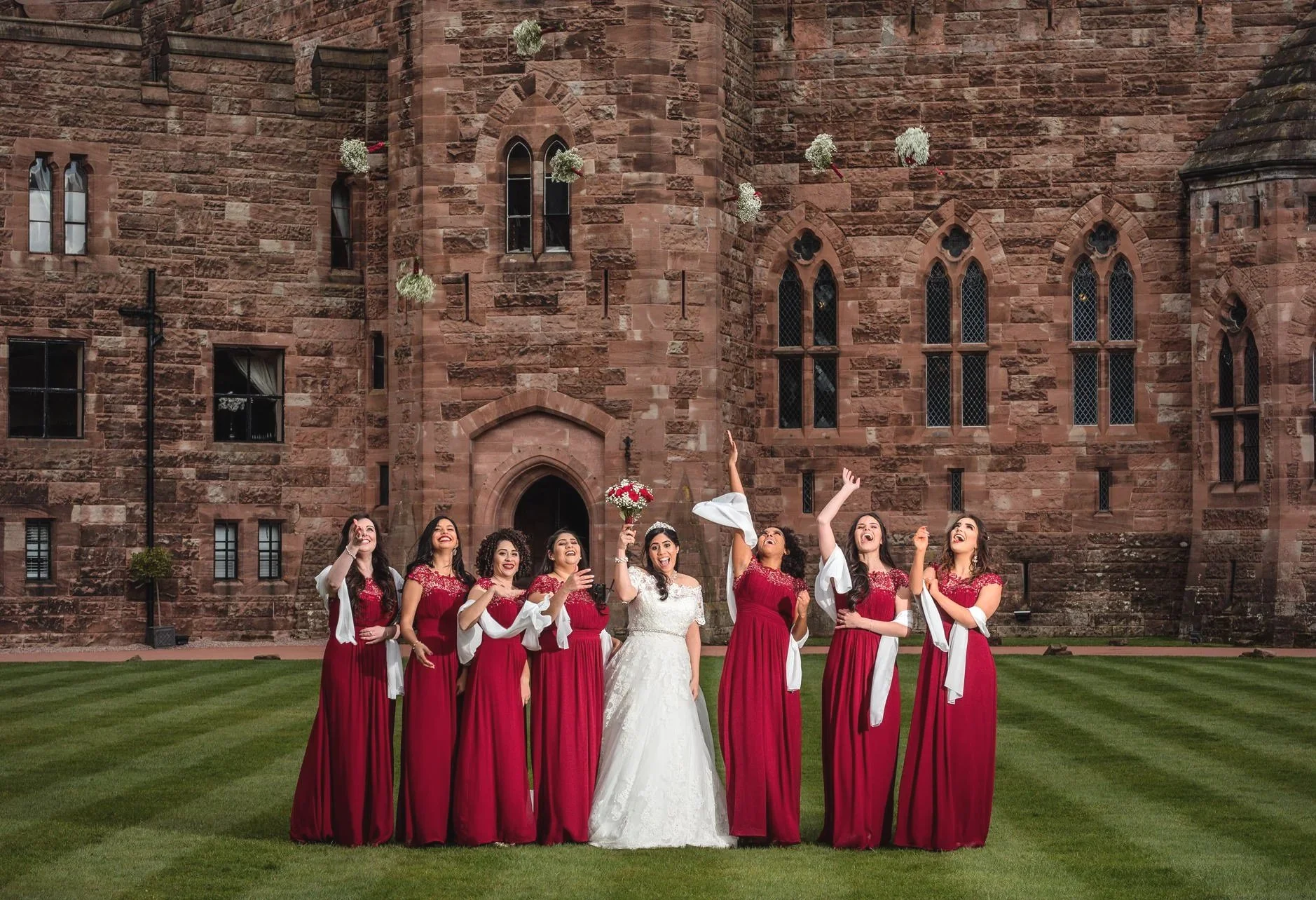 A bride and six bridesmaids in matching deep red dresses pose in front of a historic sandstone building with pointed arch windows. The bride, in a white gown, is holding a bouquet and smiling as she tosses a handful of flowers into the air. The bridesmaids are laughing and looking up, enjoying the moment.