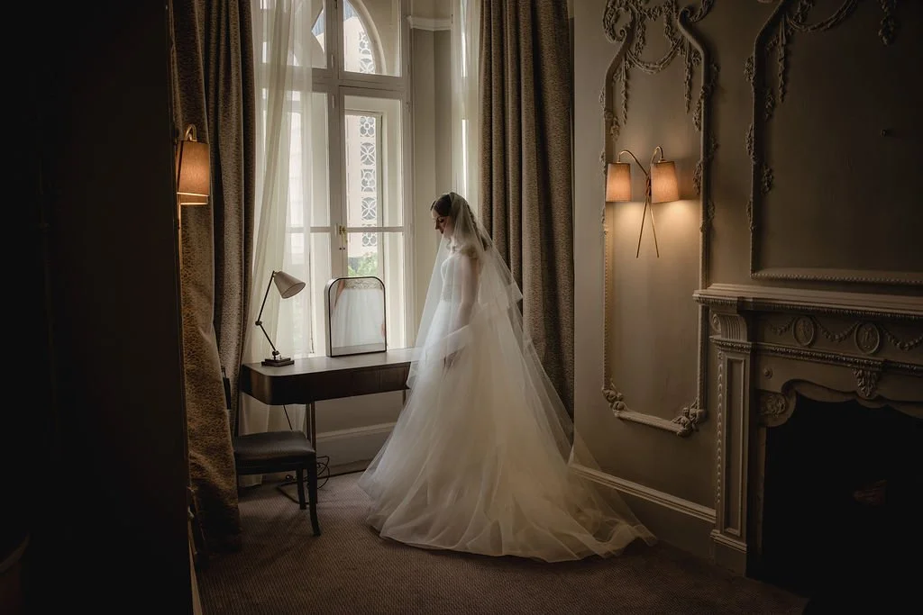 Bride in a wedding dress standing by a window in a luxurious room with ornate decor and dim lighting.