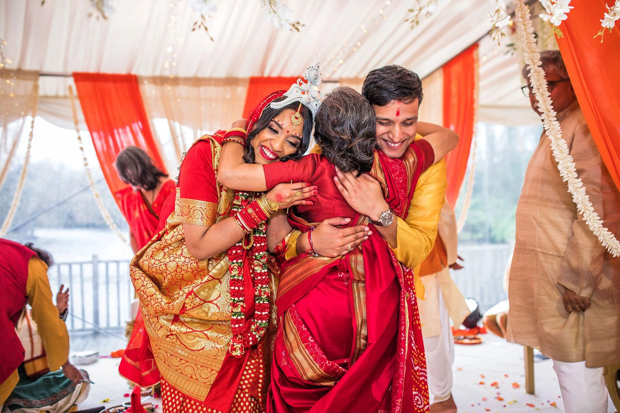 Colourful red and orange themed Asian wedding celebration bride and groom both hug the grooms mother in the ceremony room.