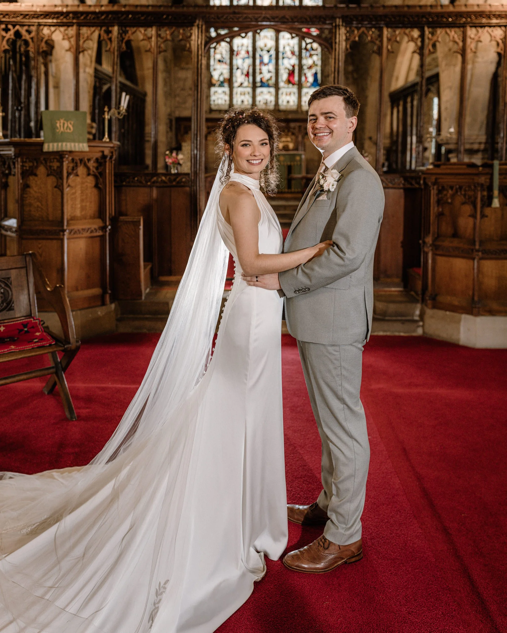 A bride and groom standing inside a church, smiling and holding each other, with stained glass windows in the background and wooden pews and red carpet on the floor.