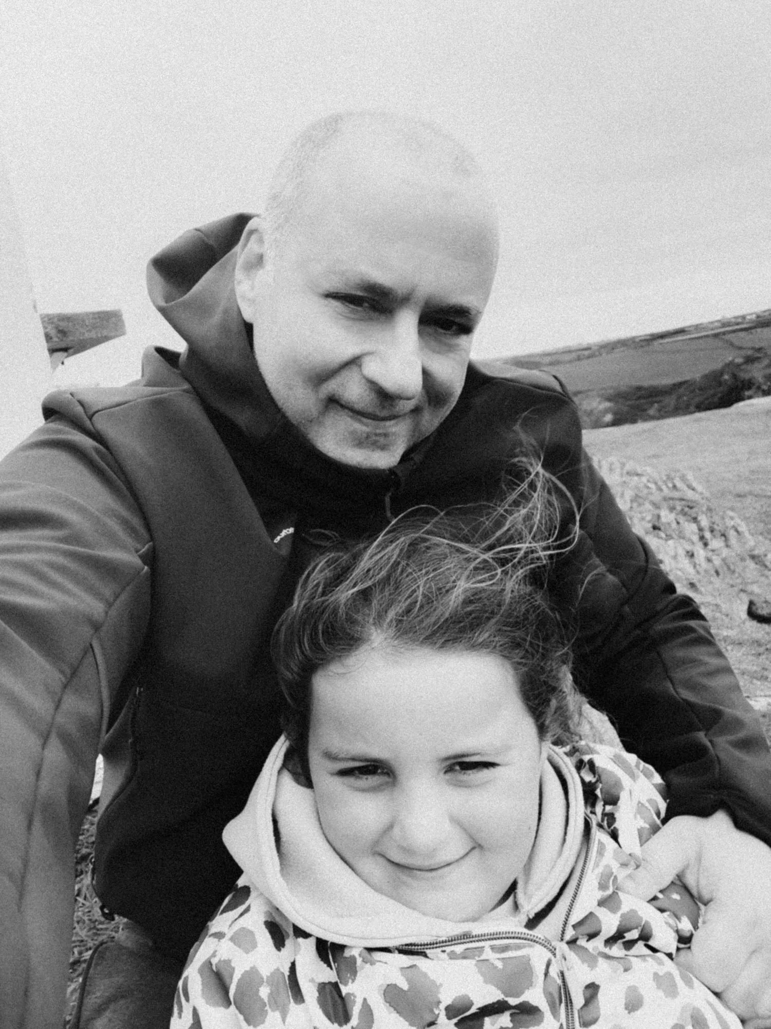 A black-and-white photo of a man and a young girl outdoors in a rural landscape. The man has a close-shaved haircut and is wearing a jacket with a hoodie, smiling slightly. The girl has curly hair and is smiling, wearing a patterned jacket with a hoodie.