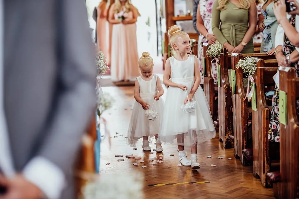 Two young girls in white dresses walking down the aisle at a wedding, carrying small baskets, with guests seated on either side.