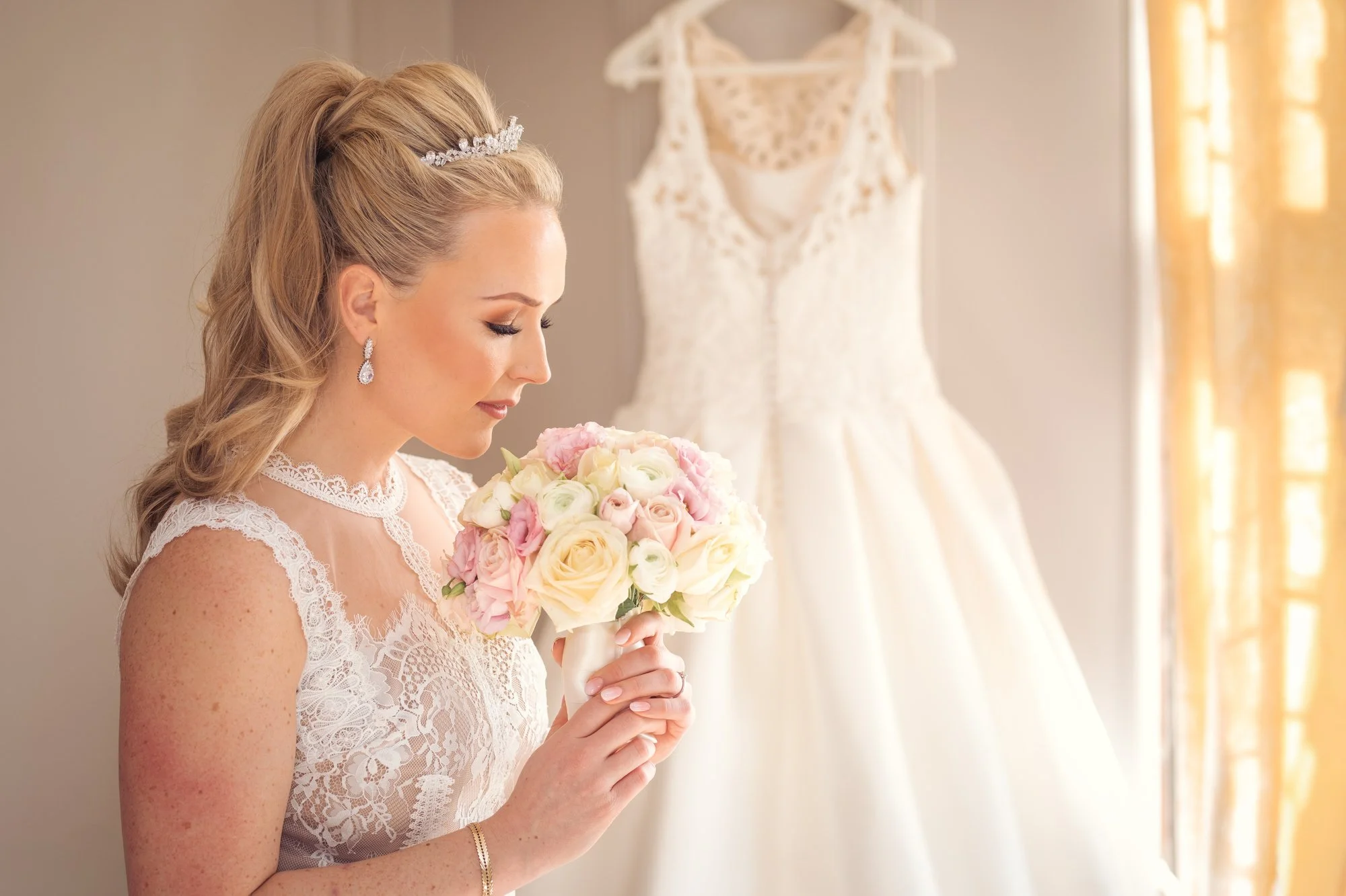 A bride with blonde hair styled in loose curls, wearing a lace wedding dress and jewelry, holding a bouquet of pink and white roses. Behind her, a wedding dress hangs on the wall, and sunlight streams through the window.