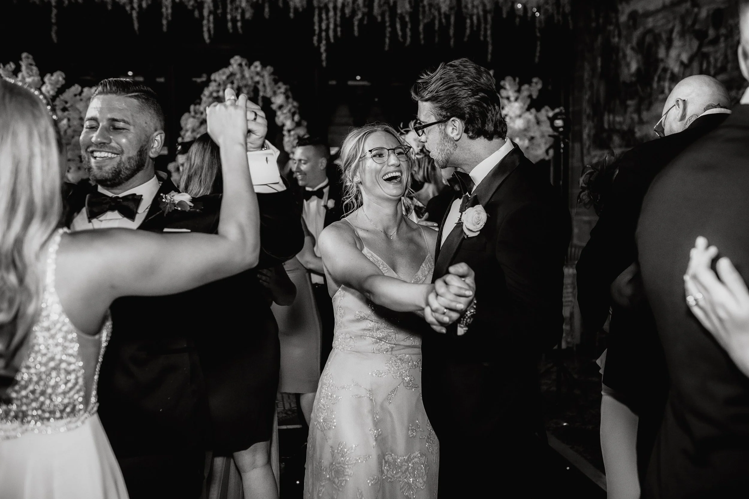 Black and white photo of a wedding reception dance floor, with a bride and groom dancing and smiling, surrounded by happy guests dancing and celebrating.