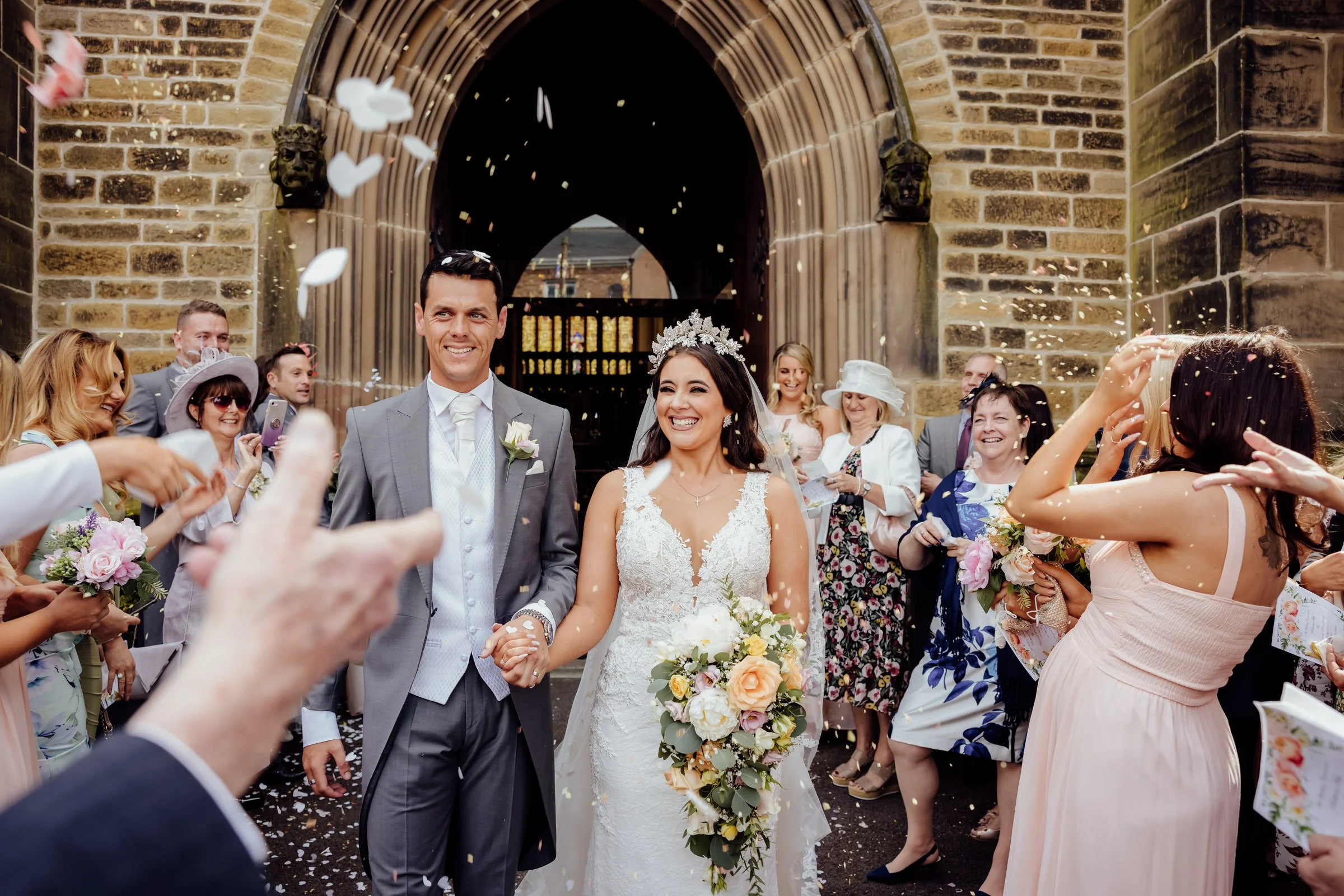 Bride and groom walking out of a church while celebrating with guests throwing confetti, with a brick church entrance in the background.