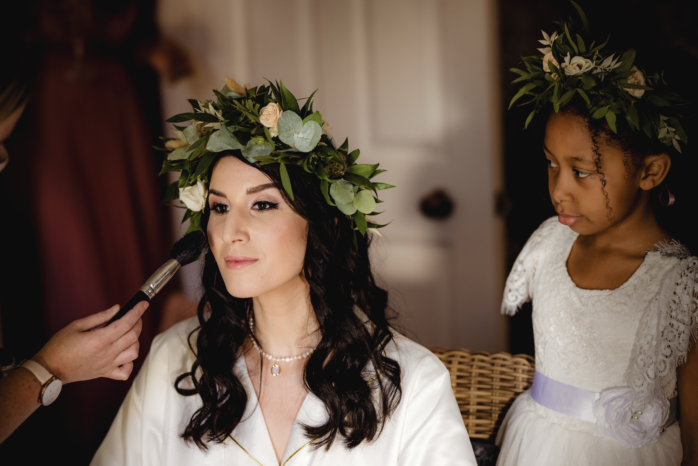 Woman with dark hair and light skin having makeup applied, wearing a crown of greenery and white flowers, with a young girl with dark curly hair and dark skin watching, also wearing a crown of greenery and white flowers, in an indoor setting.
