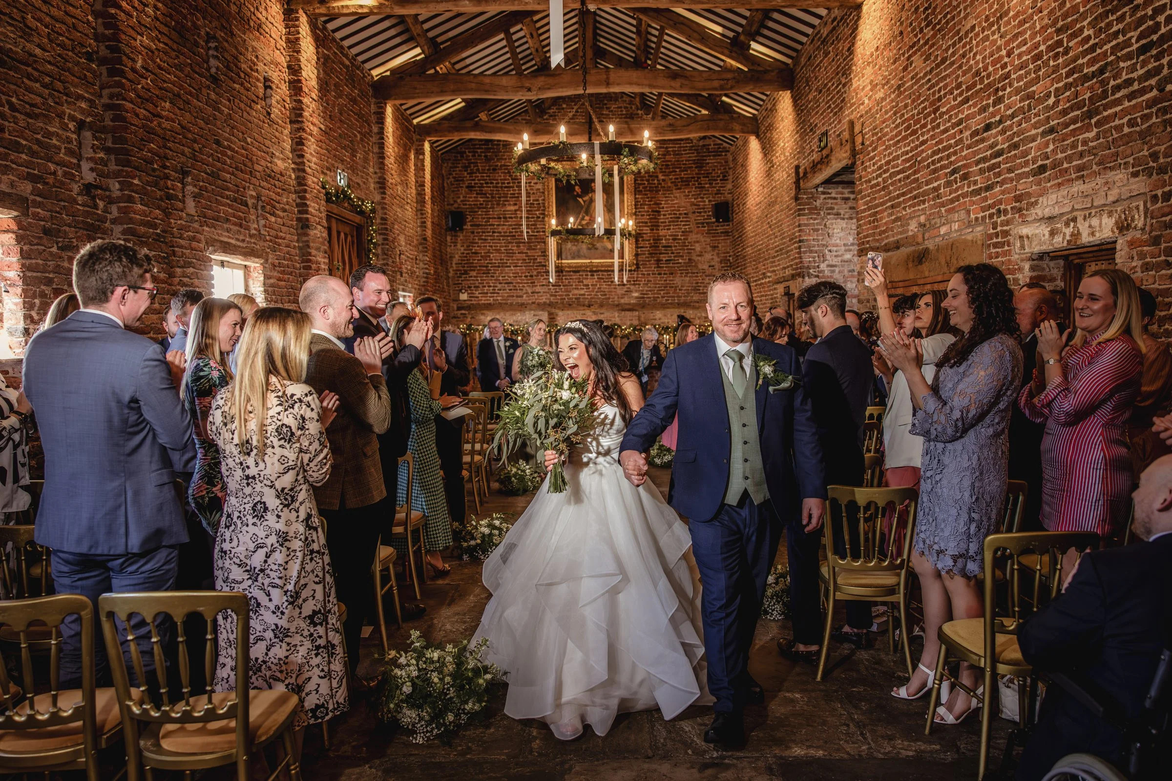 A wedding couple walking down the aisle in a brick church, holding hands, with guests clapping and smiling around them.
