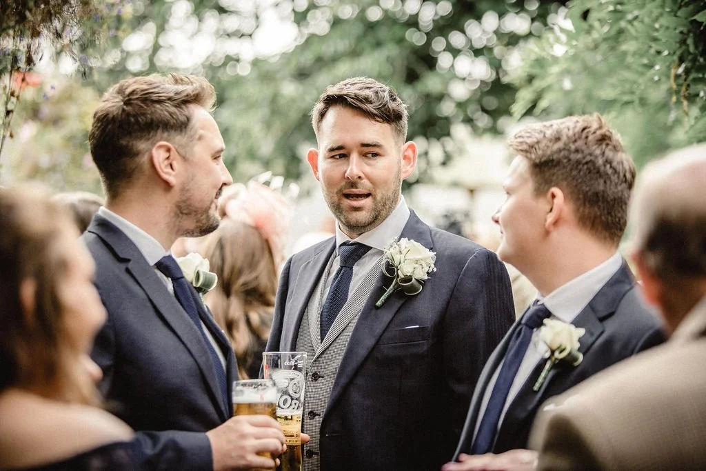 Group of men in suits at outdoor wedding reception, one holding a beer, engaged in conversation.