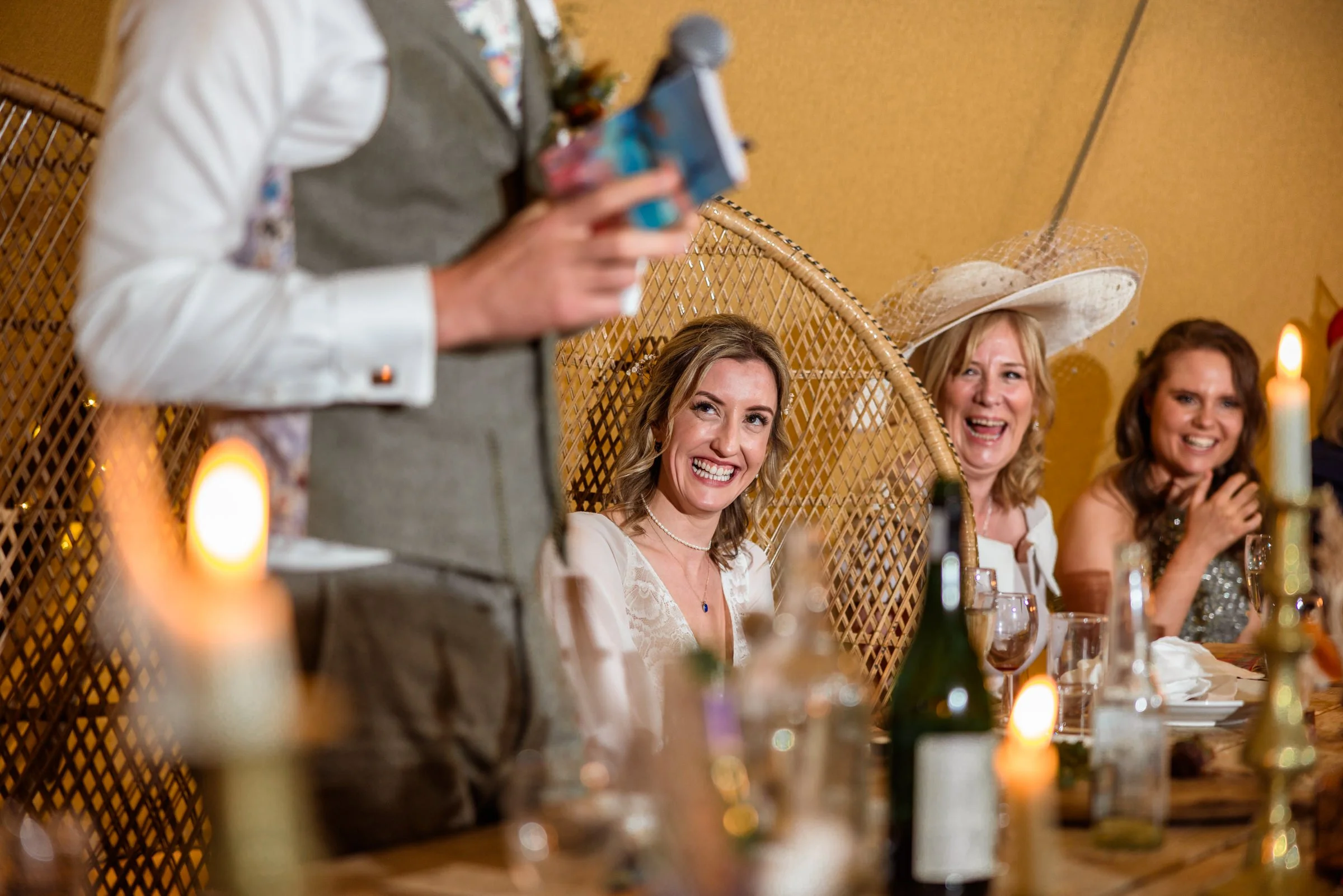 Women at a celebration sitting at a table, smiling and laughing, with candles and wine glasses, and a man standing speaking.
