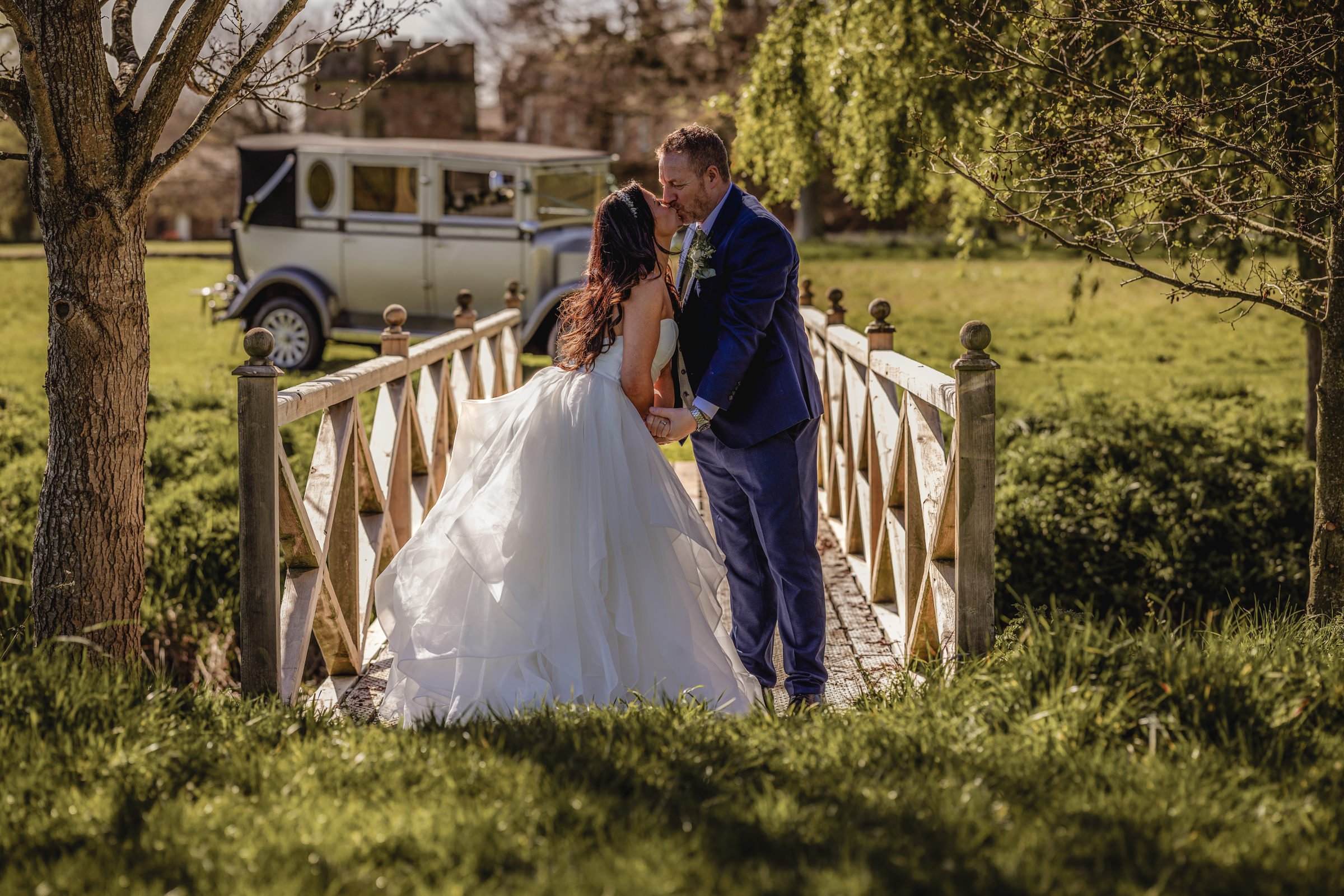 A bride and groom kissing on a small wooden bridge in a park, with a vintage car in the background and trees surrounding them.