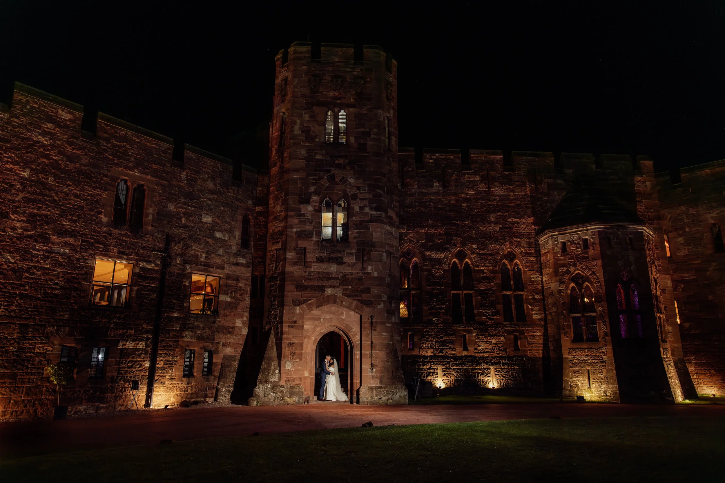 A bride and groom embracing in front of a castle at night, illuminated by exterior lights, with a dark sky above.