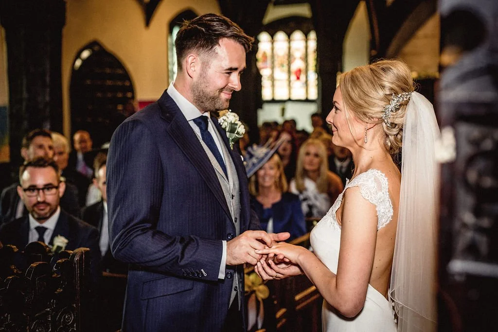 A bride and groom exchange rings during their wedding ceremony inside a church, with guests watching in the background.