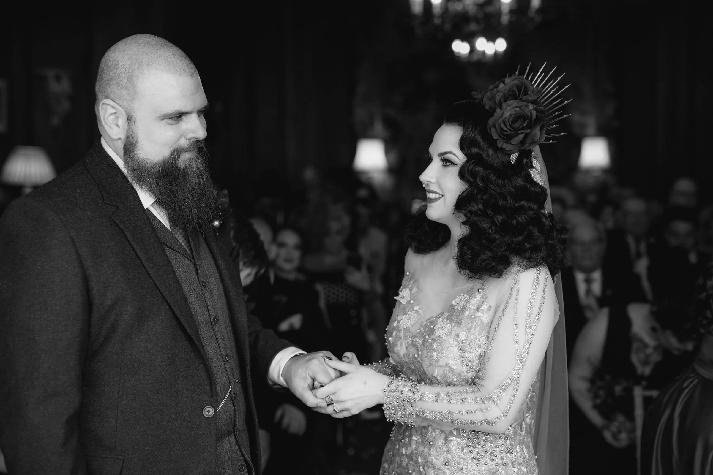 Black and white photo of a bride and groom exchanging vows, holding hands, during their wedding ceremony, with guests seated in the background.
