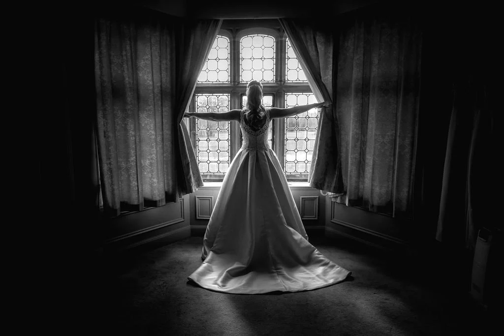 A woman in a wedding gown standing by a large window with curtains, holding the curtains open with her arms outstretched.