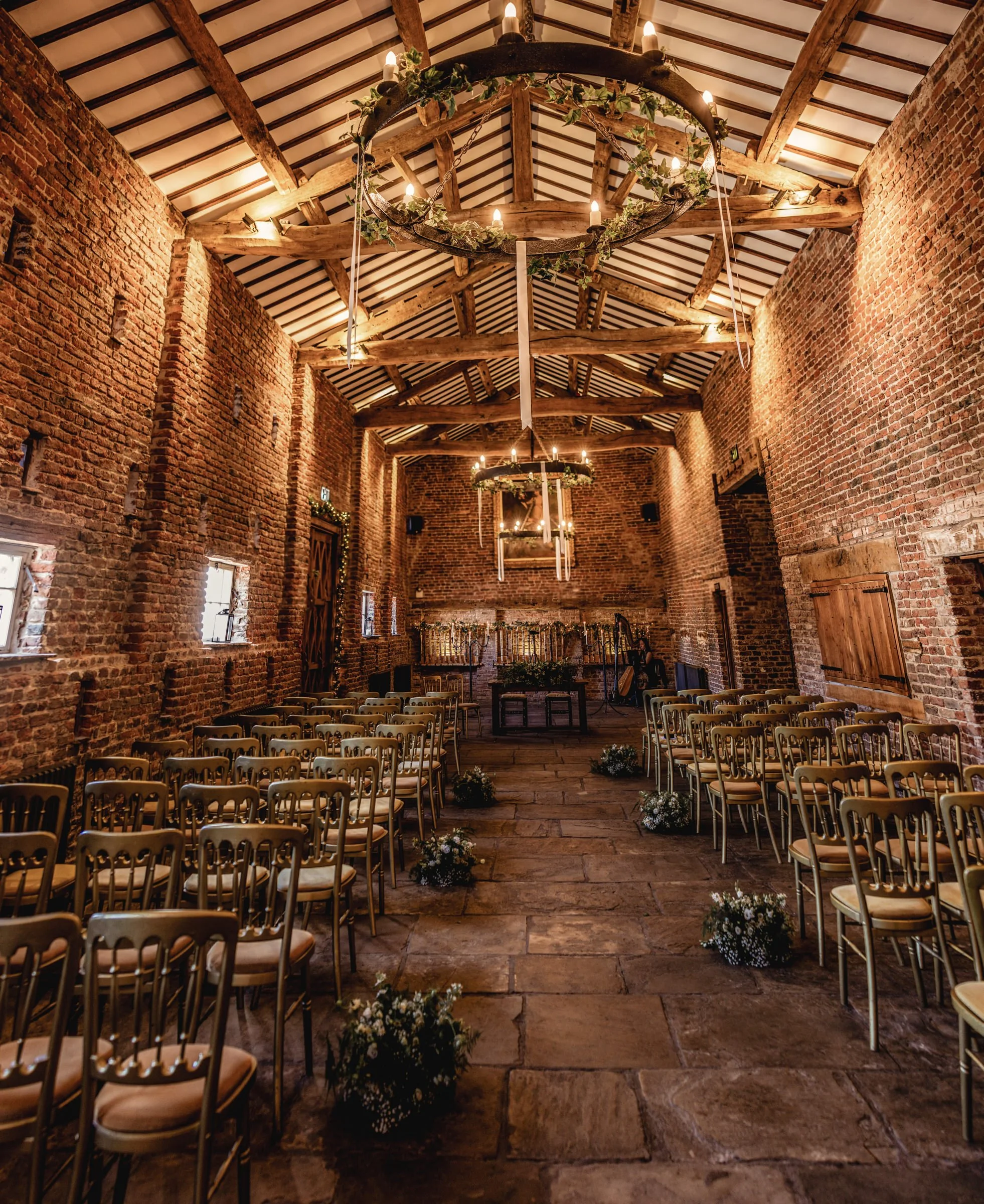 Interior of a rustic wedding venue with wooden beams, brick walls, and a stone floor. Rows of chairs are set for a ceremony, decorated with small flower arrangements on the aisle. Hanging chandeliers with greenery and candle-like lights are suspended from the ceiling.