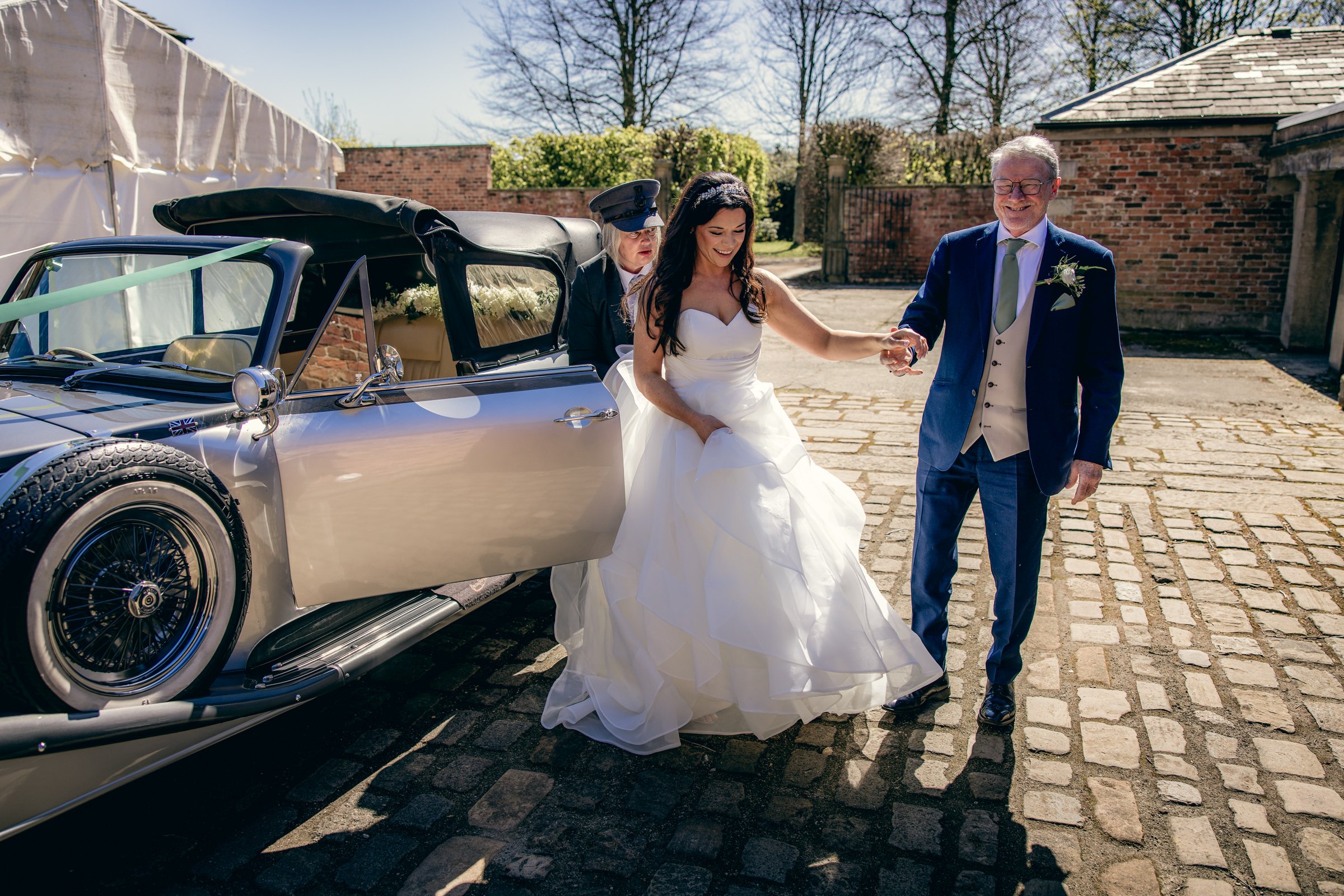 A bride, groom, and a woman, possibly the wedding officiant, exit a silver vintage car on a cobblestone street on a sunny day, with a brick wall and trees in the background.