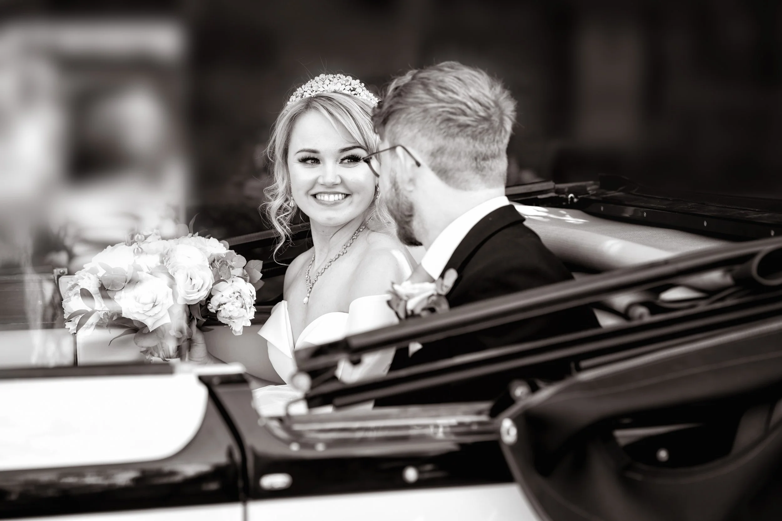 Black-and-white photo of a bride and groom sitting in a car, smiling at each other. The bride is holding a bouquet of flowers and wearing a tiara, necklace, and strapless dress. The groom is wearing glasses and a tuxedo with a boutonniere.