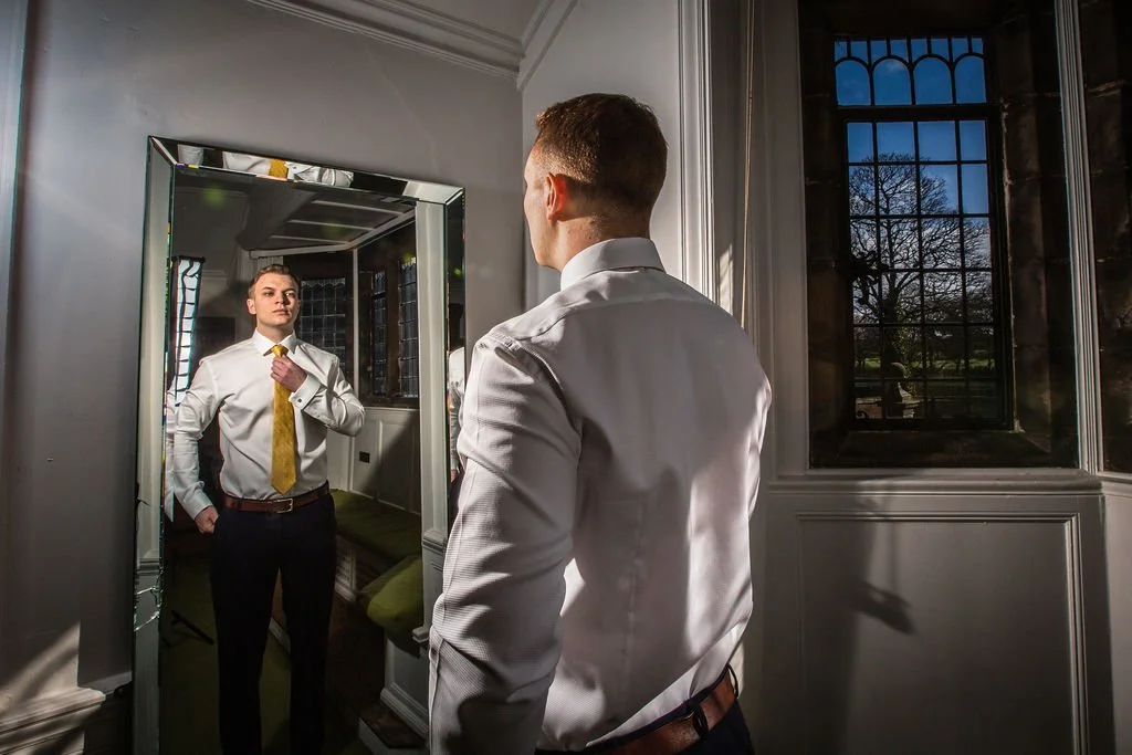 A man in a white dress shirt and yellow tie looking at himself in a full-length mirror, adjusting his tie.