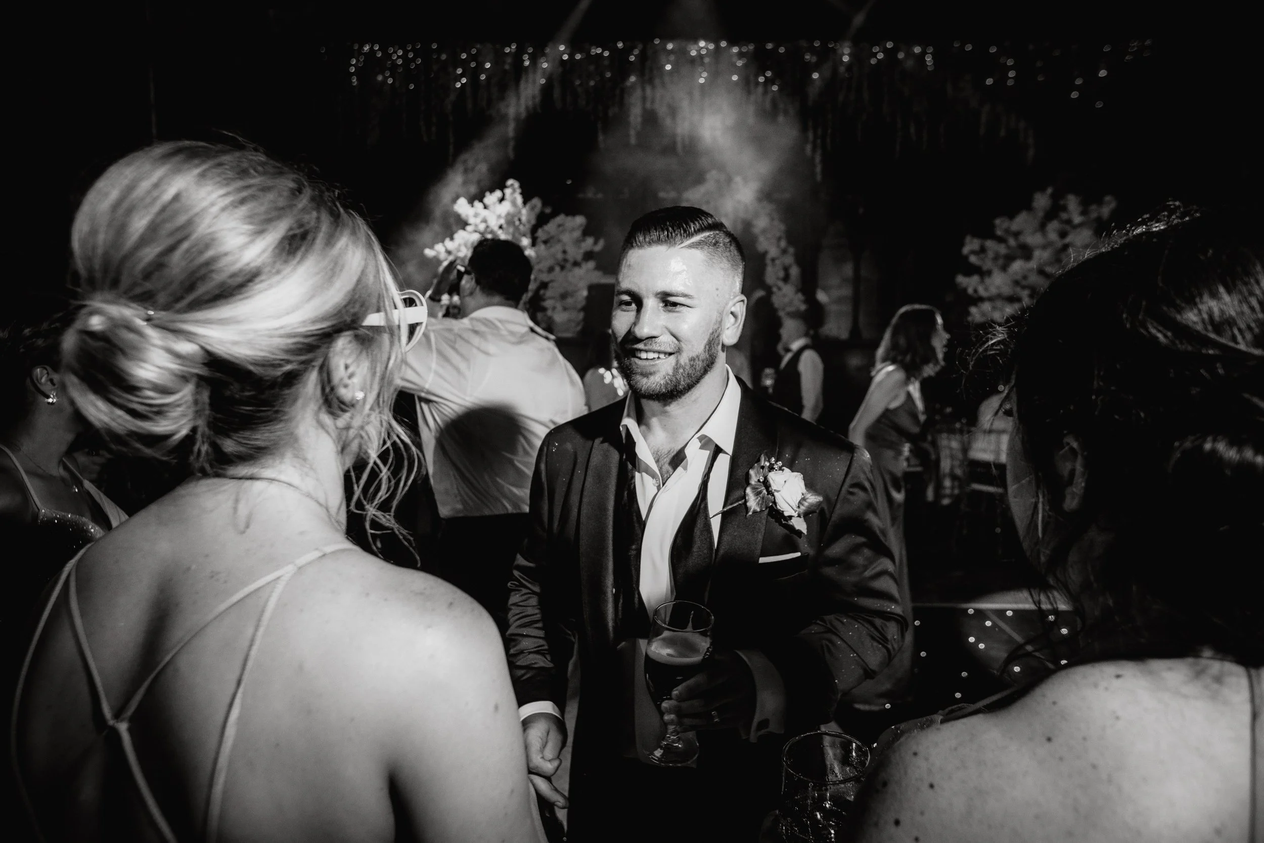 A black and white photo of a man in a suit smiling and holding a drink, engaging in conversation at a social event with women around him.