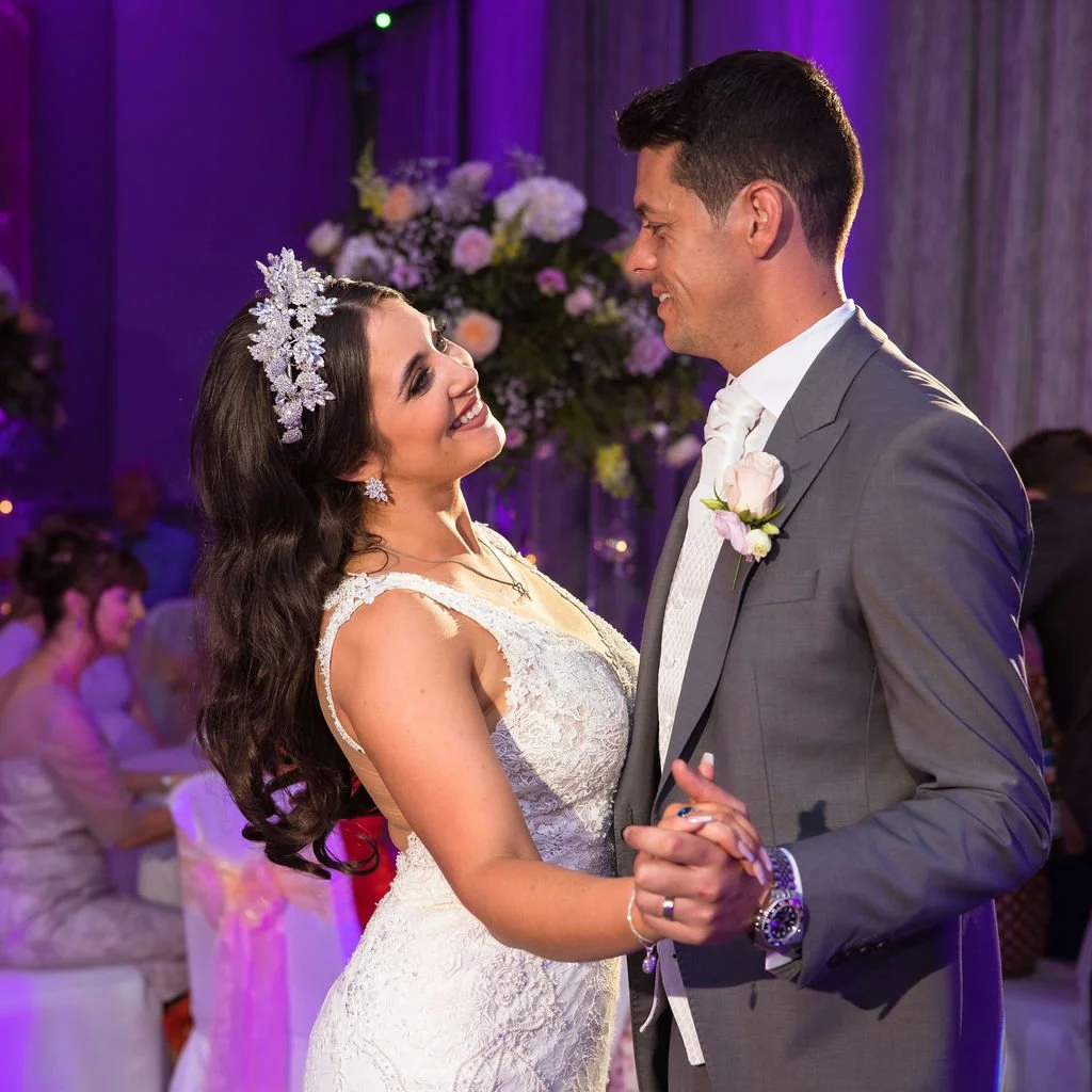 A bride and groom dancing at their wedding reception, smiling at each other with a background of purple lighting and floral decorations.