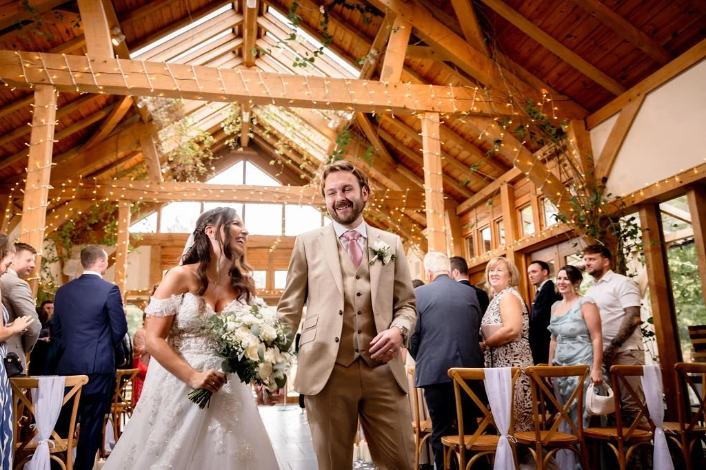 Wedding reception inside a charming wooden barn with string lights and greenery, featuring a smiling bride holding a bouquet, and a groom in a beige suit, surrounded by happy guests.