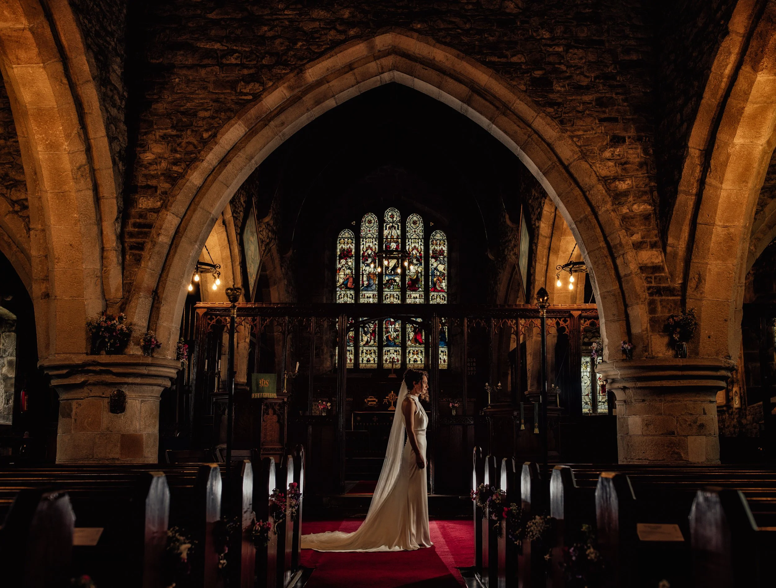 Interior of a church or chapel with a high arched ceiling, stained glass windows, and wooden pews. A woman in a white dress is standing on a red carpet in front of the altar.