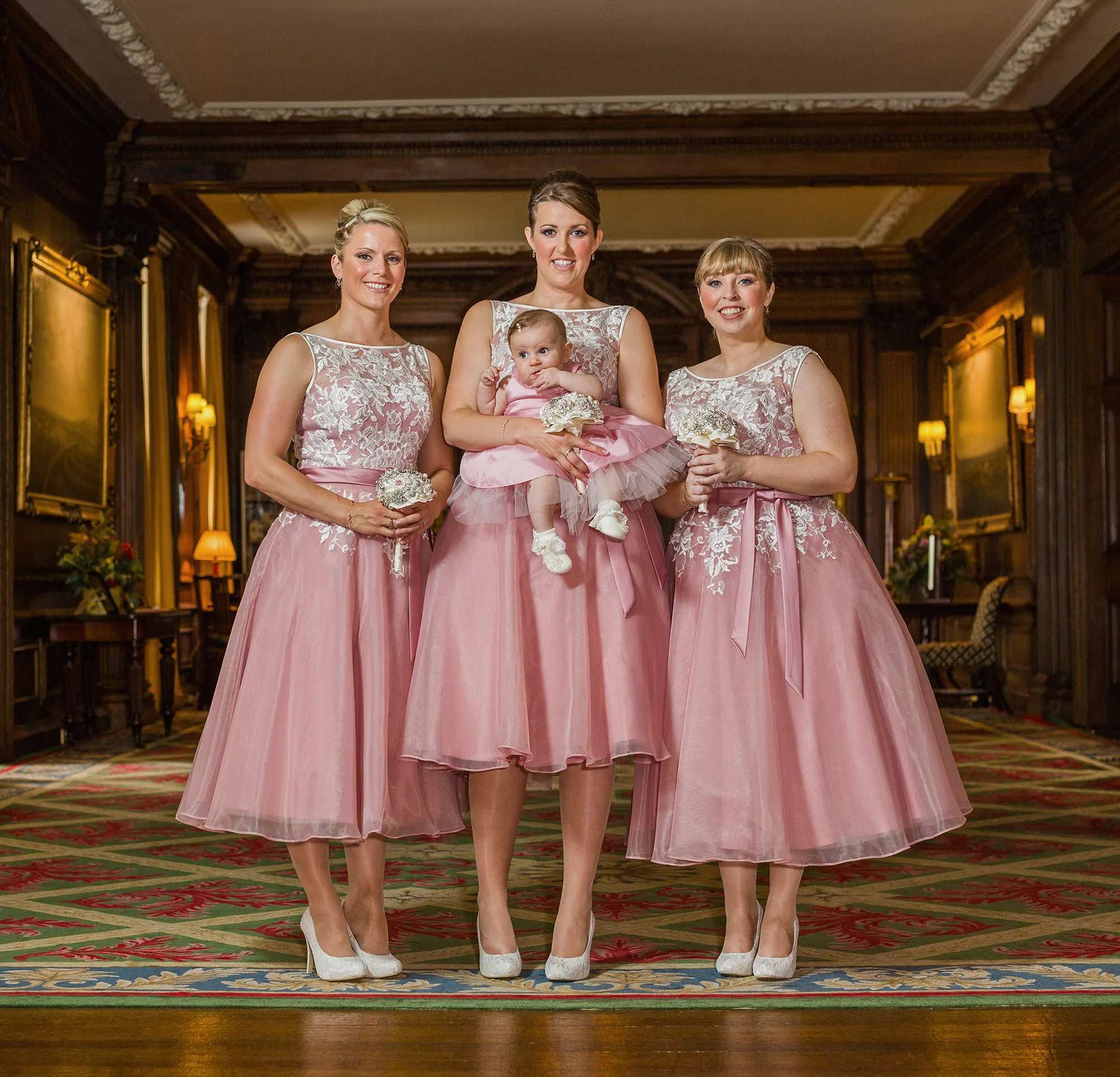 Four women and a baby in an elegant, wood-paneled room with ornate details, dressed in matching pink and white floral dresses, holding bouquets, and smiling at the camera.