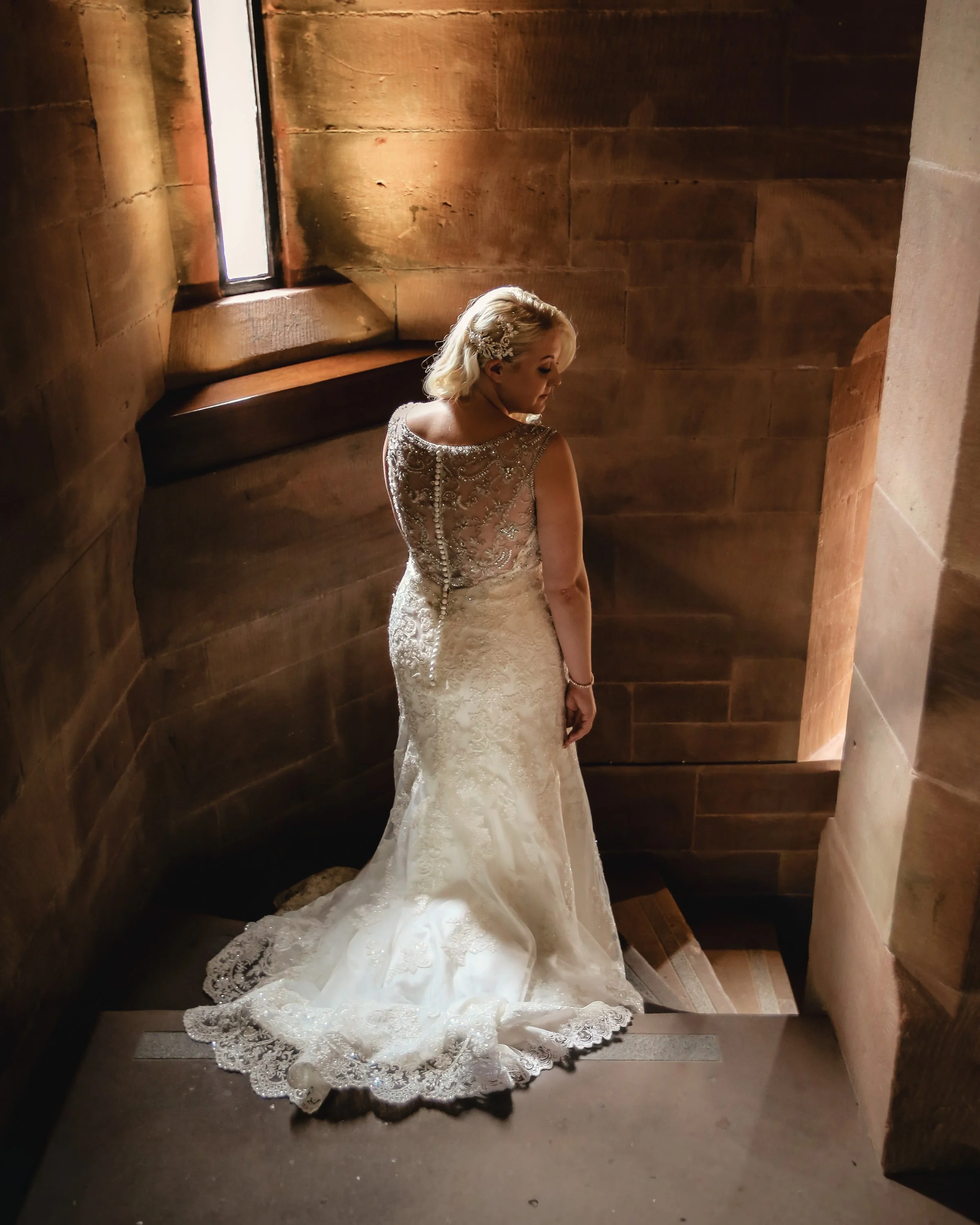 A woman in a wedding dress standing in a small, dimly lit stone room, looking downward.