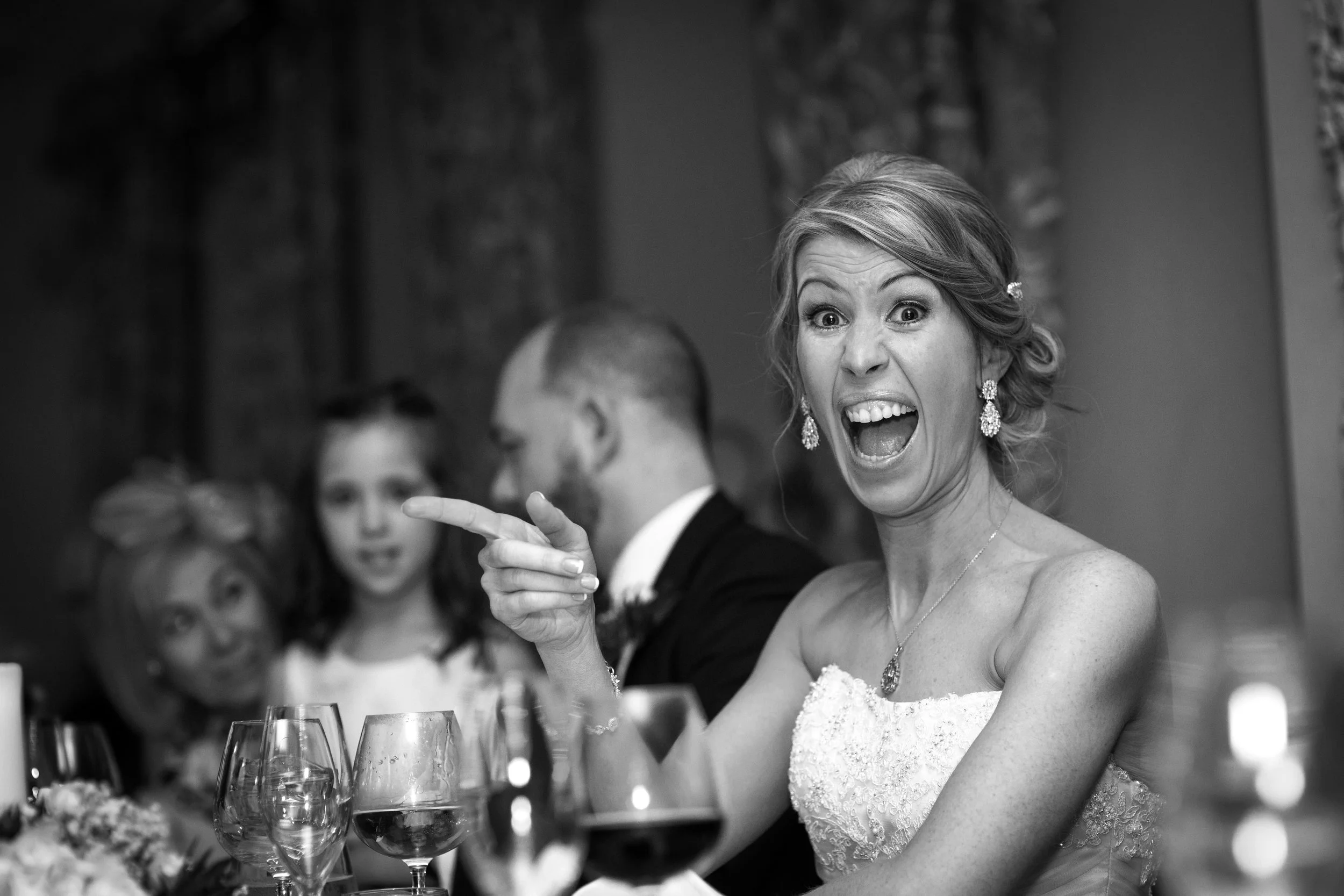 A woman in a wedding dress is smiling and talking animatedly at a wedding reception, with others in the background.