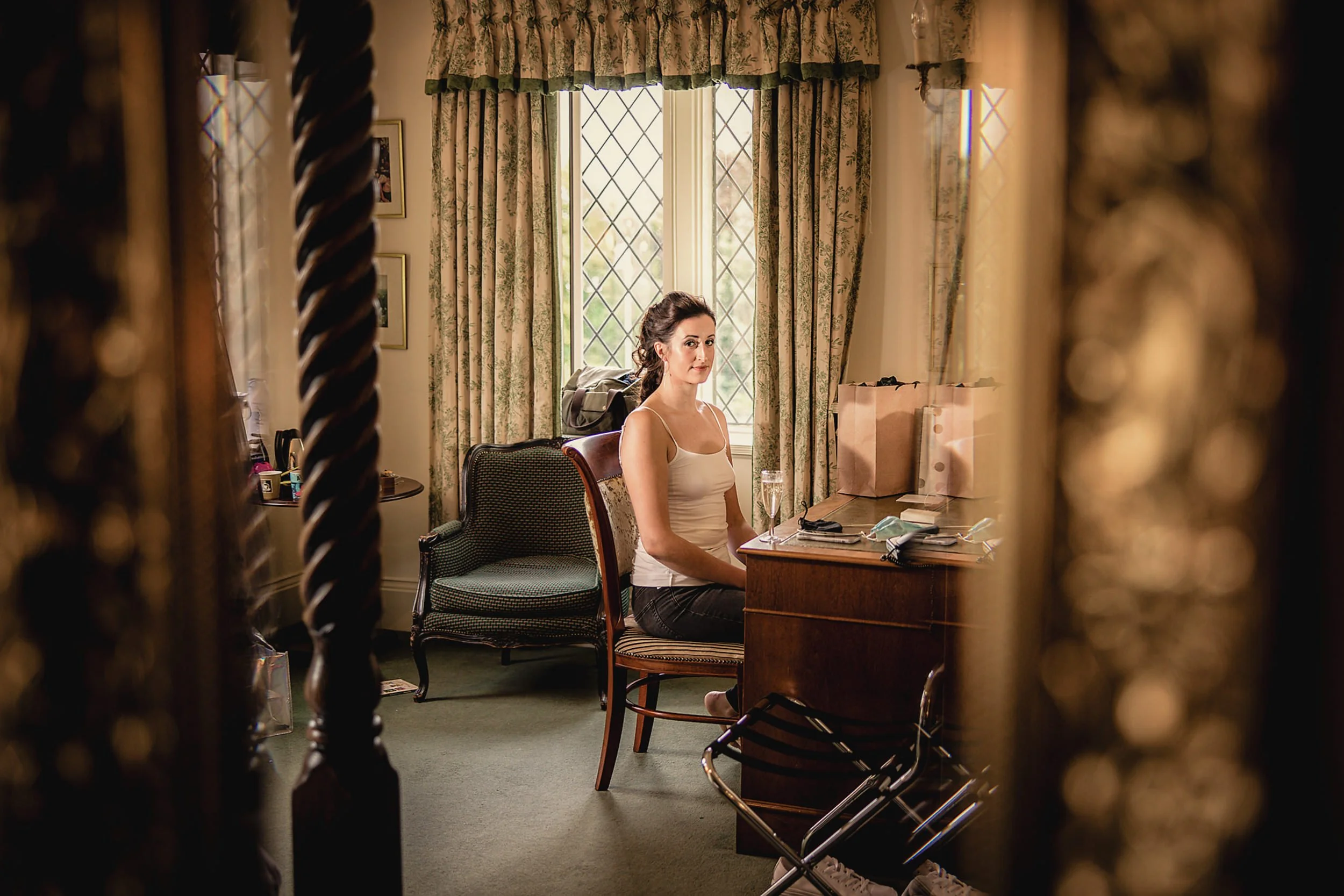 A woman with dark hair sitting at a desk in a warmly lit room, reflected in an ornate mirror, with floral curtains and a window behind her.