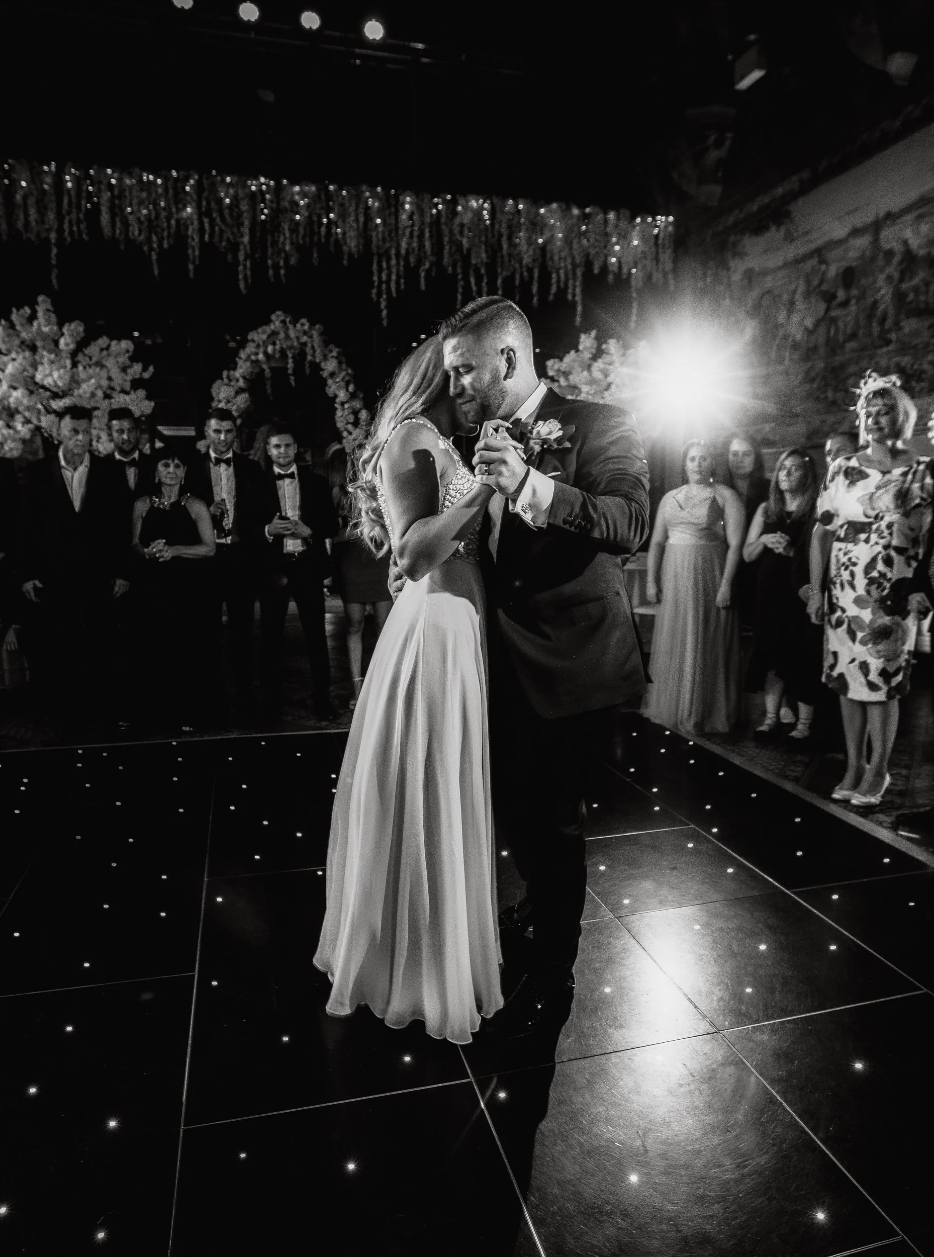 A black-and-white photo of a bride and groom sharing a dance at their wedding reception, surrounded by guests in a decorated venue.