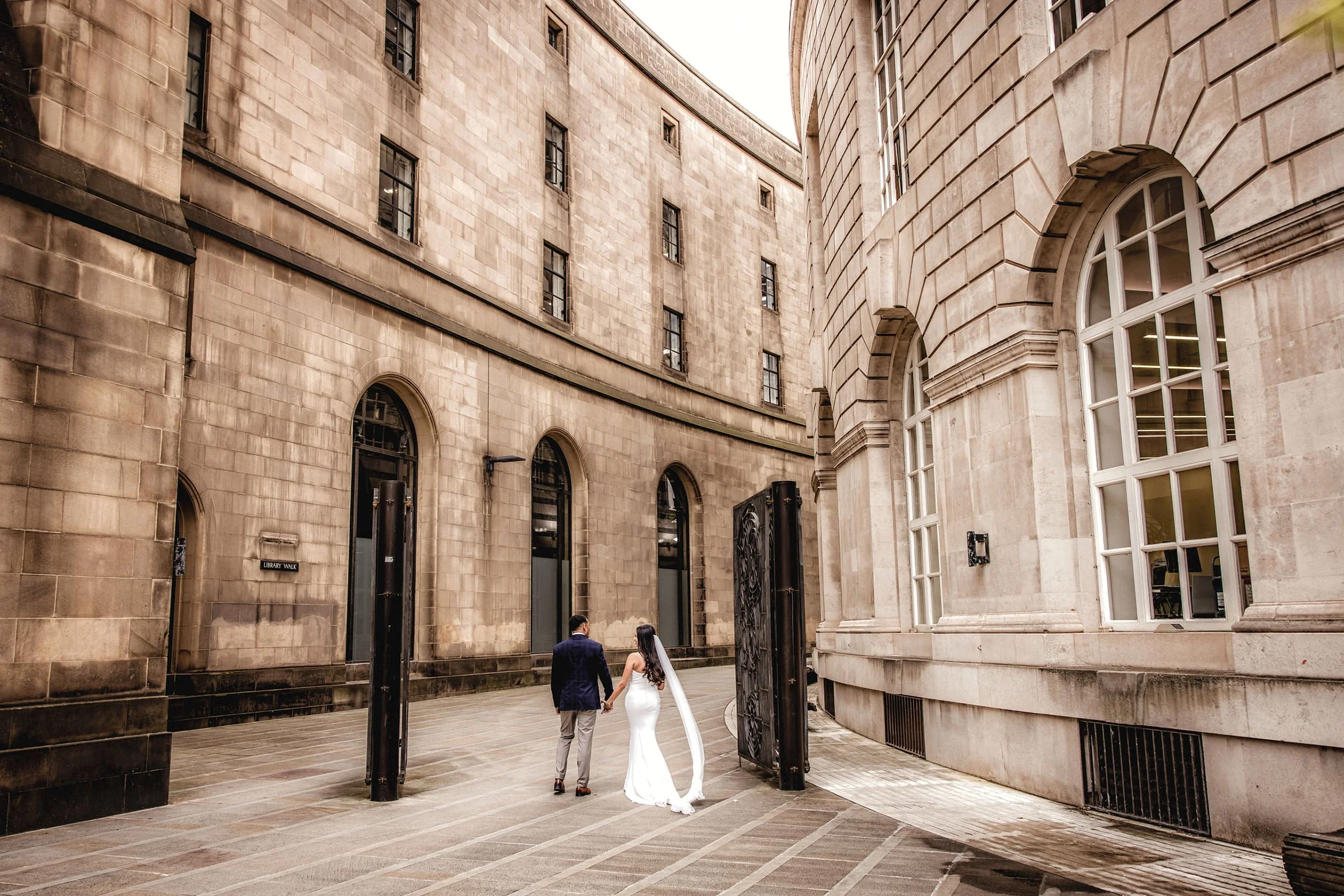 A couple walking hand-in-hand in front of large stone buildings on a city street, with the woman in a white dress and the man in a suit.