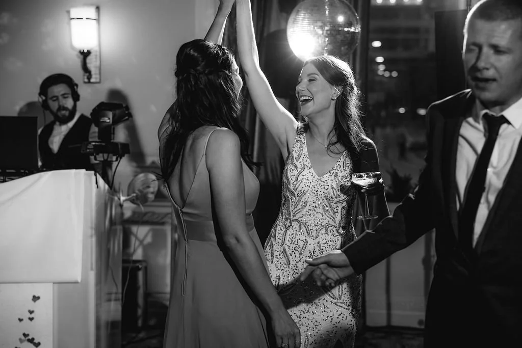 Two women dancing and smiling at a party, one woman in a patterned dress and another in a sleeveless dress, while a man in a suit shakes hands nearby. A DJ is seen in the background.