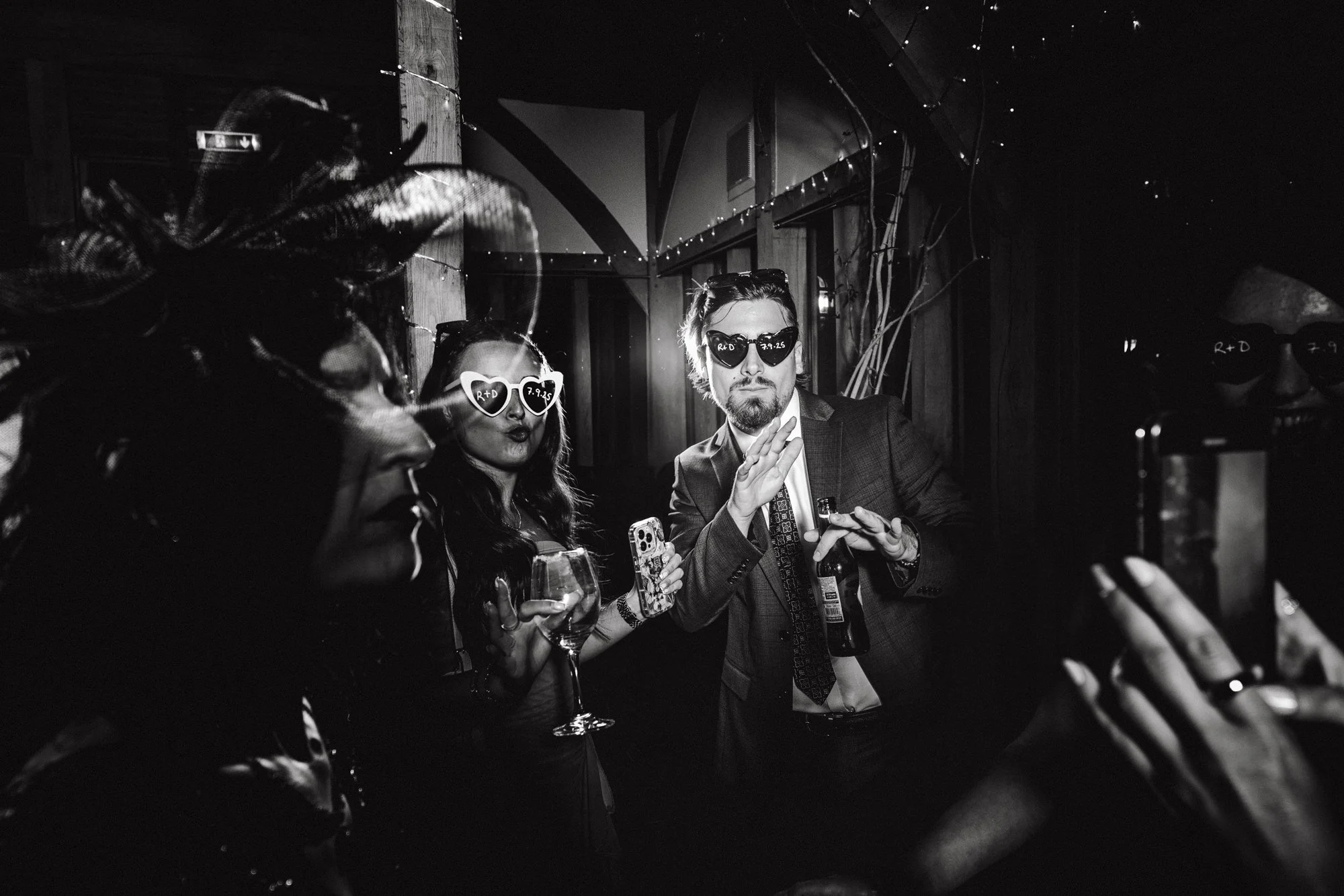 Black and white photo of three people at a party, wearing sunglasses with the words 'R+D' written on them, holding drinks, and making hand gestures.