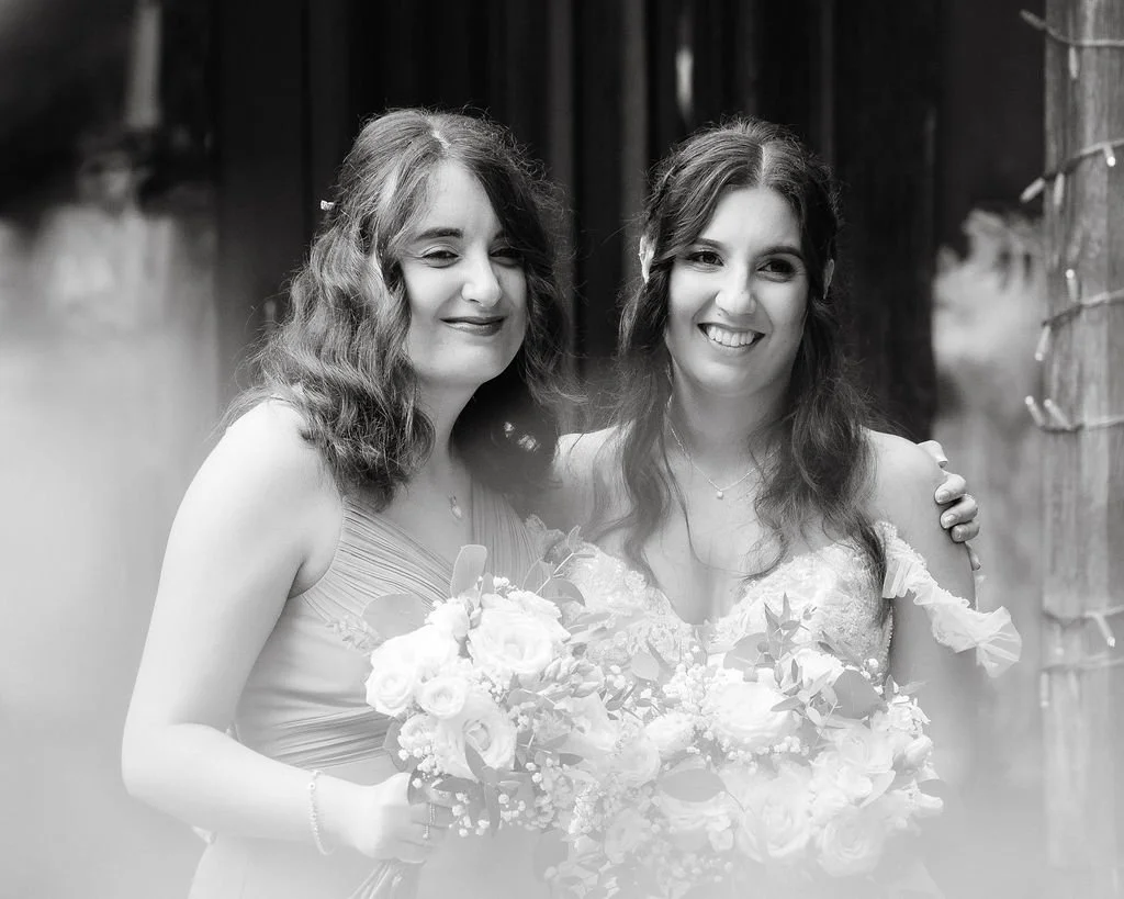Two women smiling and standing close together, one holding a bouquet of flowers, in an outdoor setting with wooden fencing in the background.
