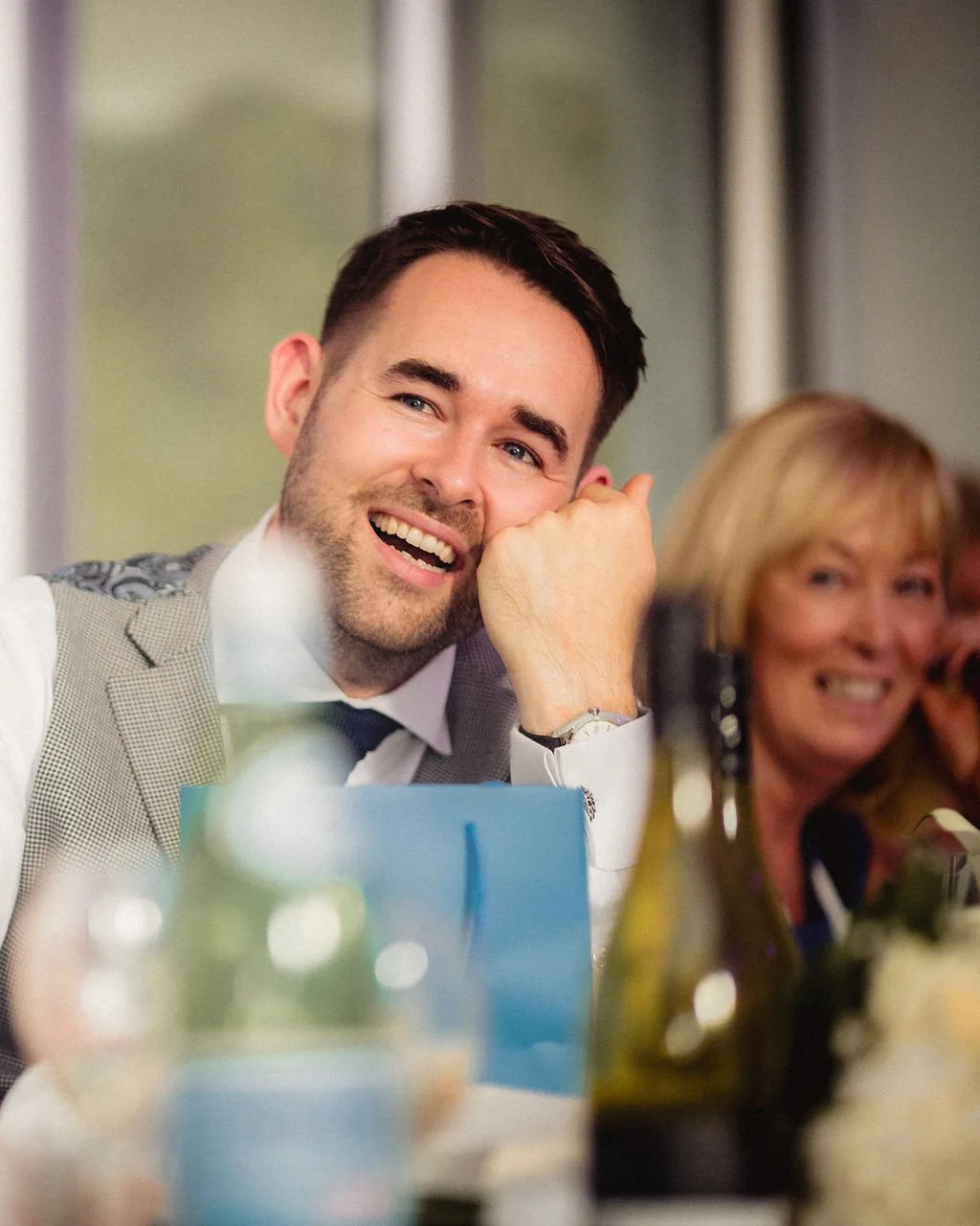 A man smiling and resting his chin on his hand at a table during a social event, with a woman smiling in the background.