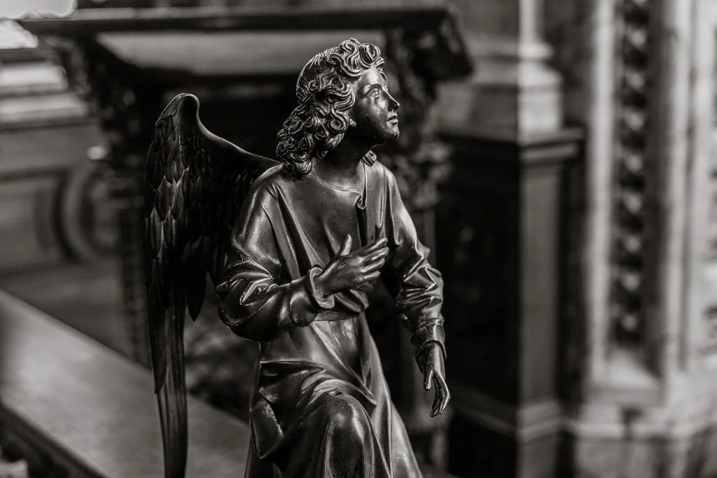 A black and white photograph of a dark-colored metal sculpture of a seated angel with large wings, curly hair, and a flowing robe, inside a building with ornate woodwork.