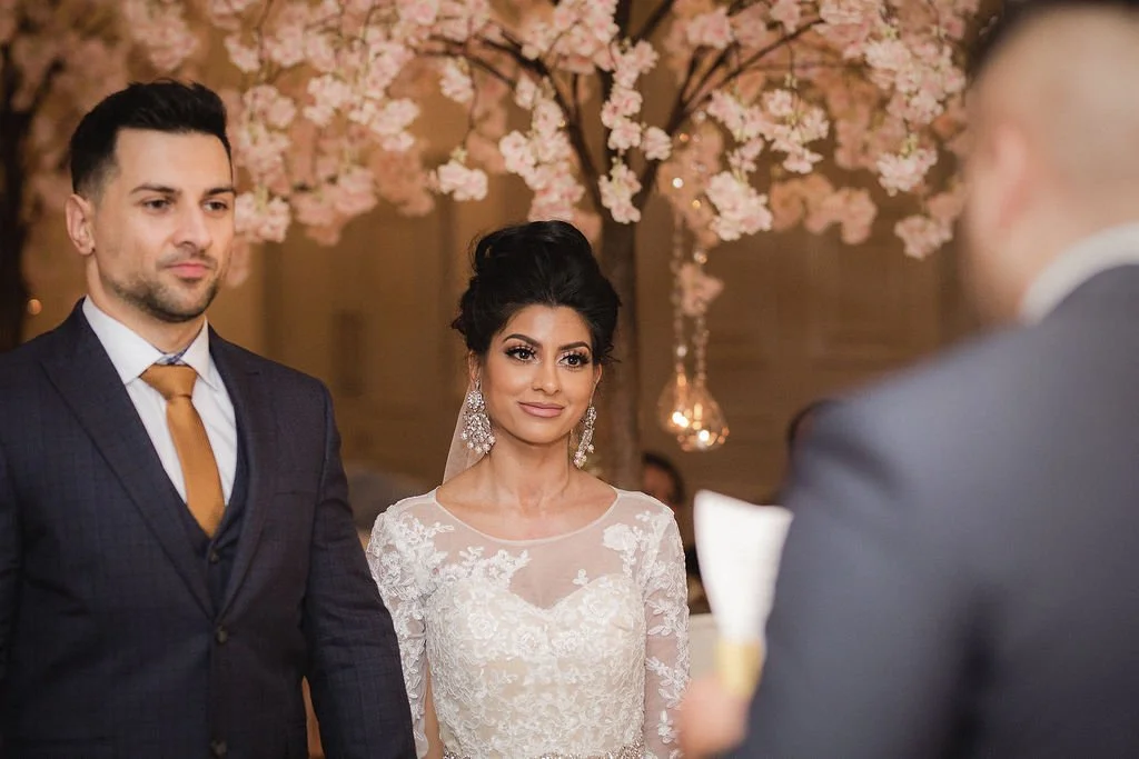 A bride and groom standing during their wedding ceremony with a person reading vows, wedding flowers, and pink cherry blossom trees in the background.