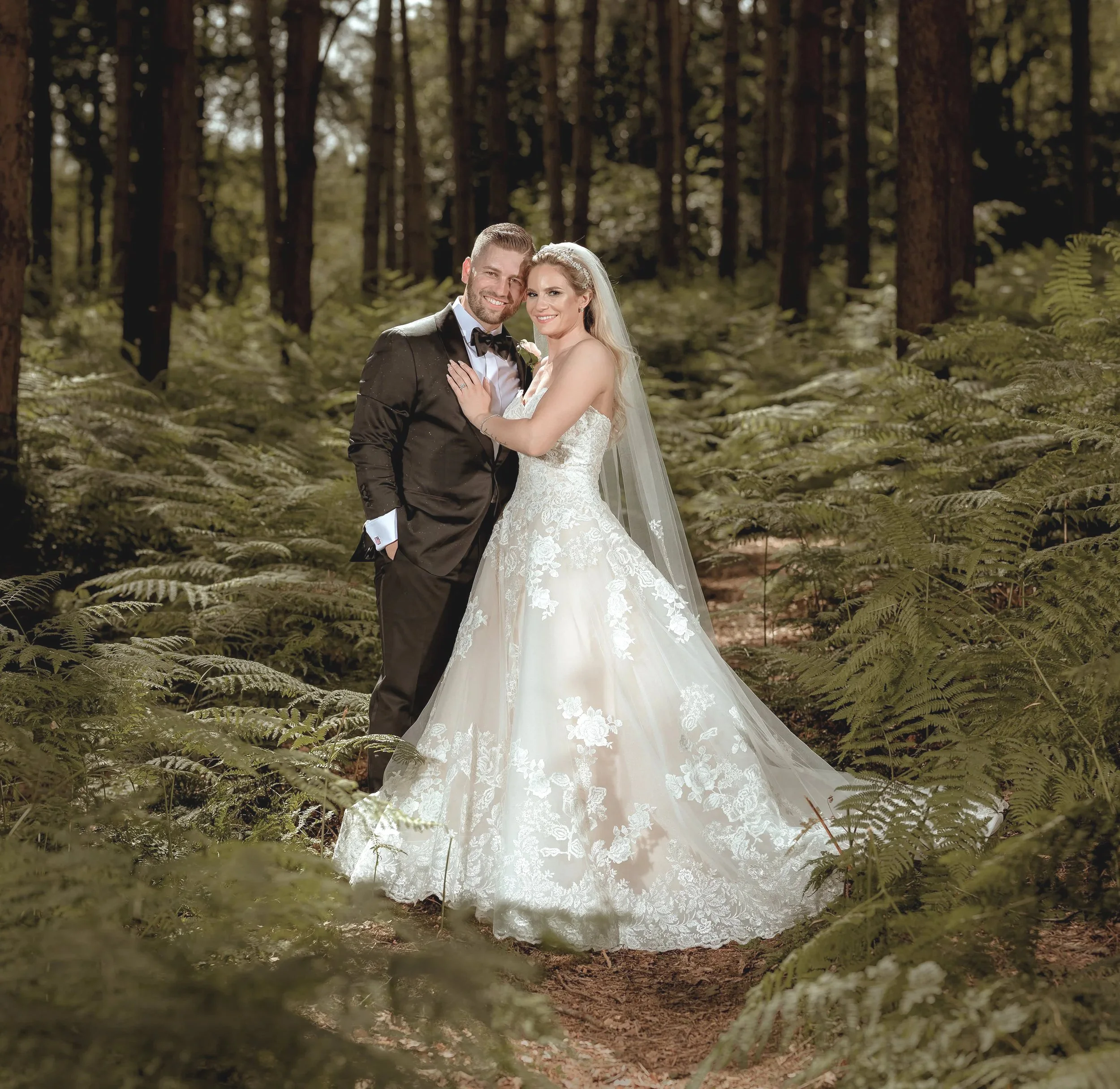A newlywed couple stands in a forest, the bride in a white lace wedding dress and veil, and the groom in a black tuxedo with a bow tie, smiling and posing together.