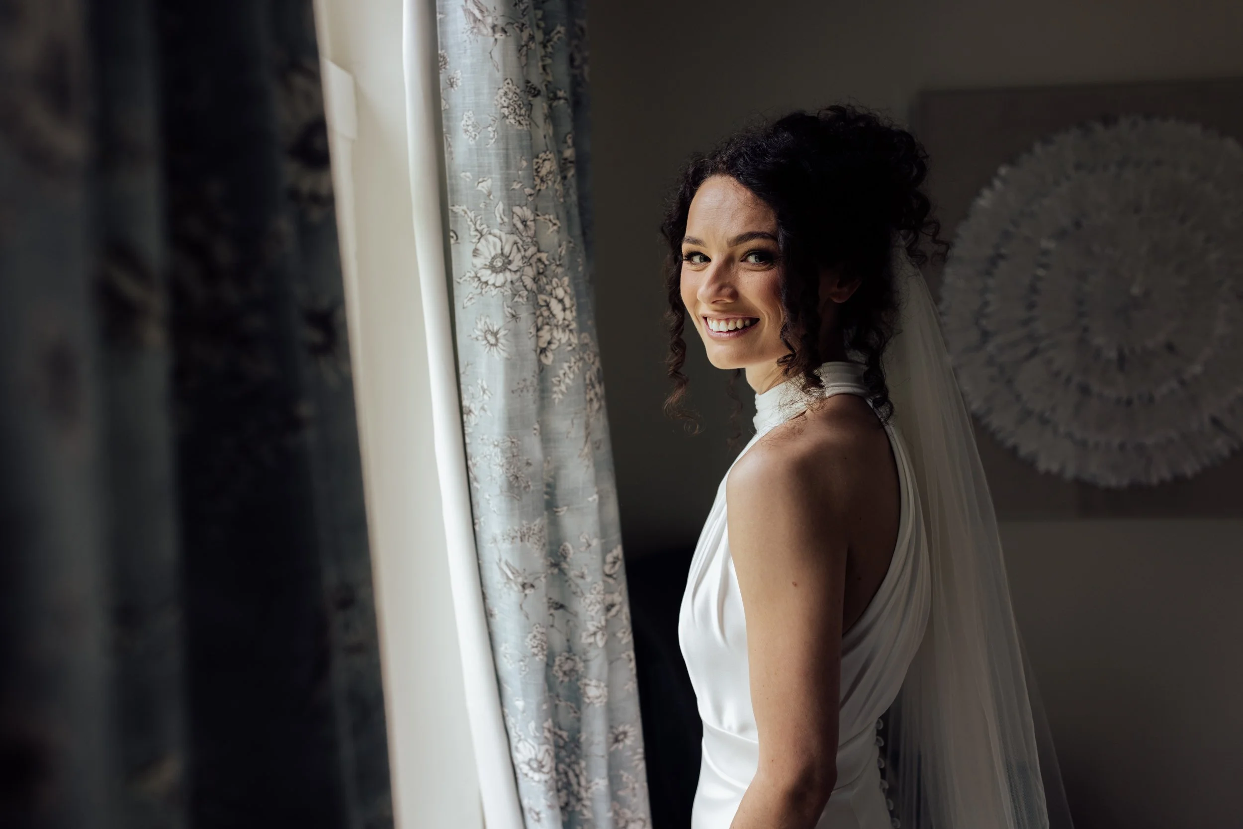 A woman with dark curly hair smiles while looking out a window, wearing a white sleeveless dress, with curtains and wall art visible in the background.