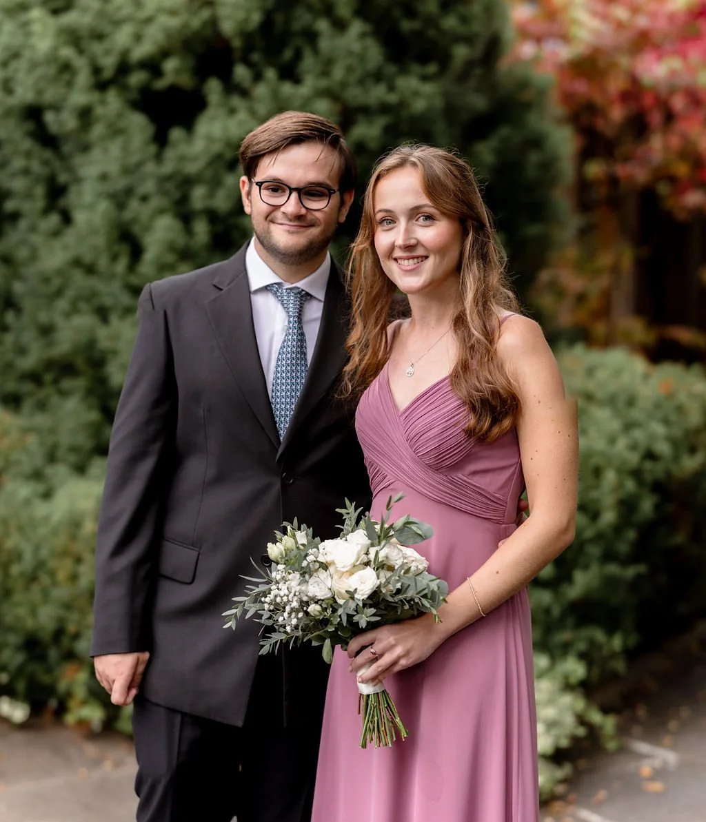 A young man and woman smiling outdoors, woman holding a bouquet of flowers, woman wearing a pink dress, man in a suit and tie.