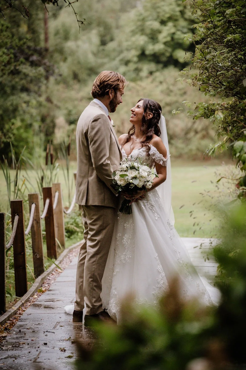 A bride and groom standing on a wooden pathway in a lush garden, gazing at each other, with the bride holding a bouquet of white flowers.