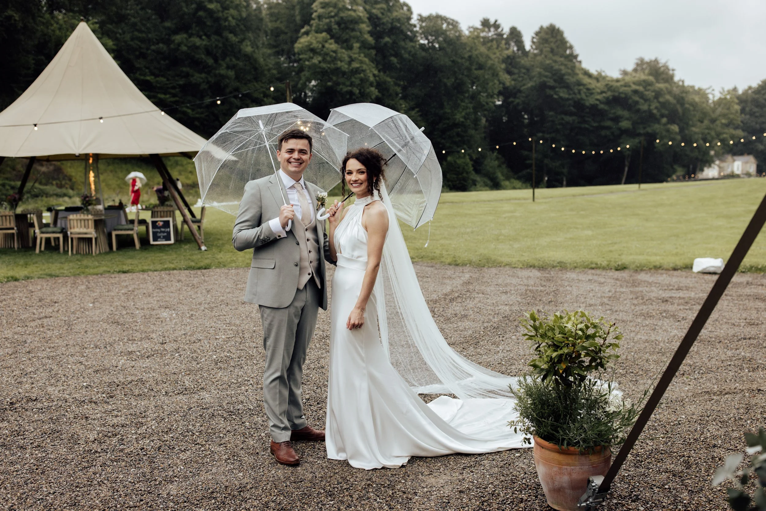 A newlywed couple stands outdoors holding clear umbrellas during a light rain. The groom wears a light gray suit with a white shirt and beige tie, and the bride wears a white wedding gown with lace details. They are smiling. In the background, there 