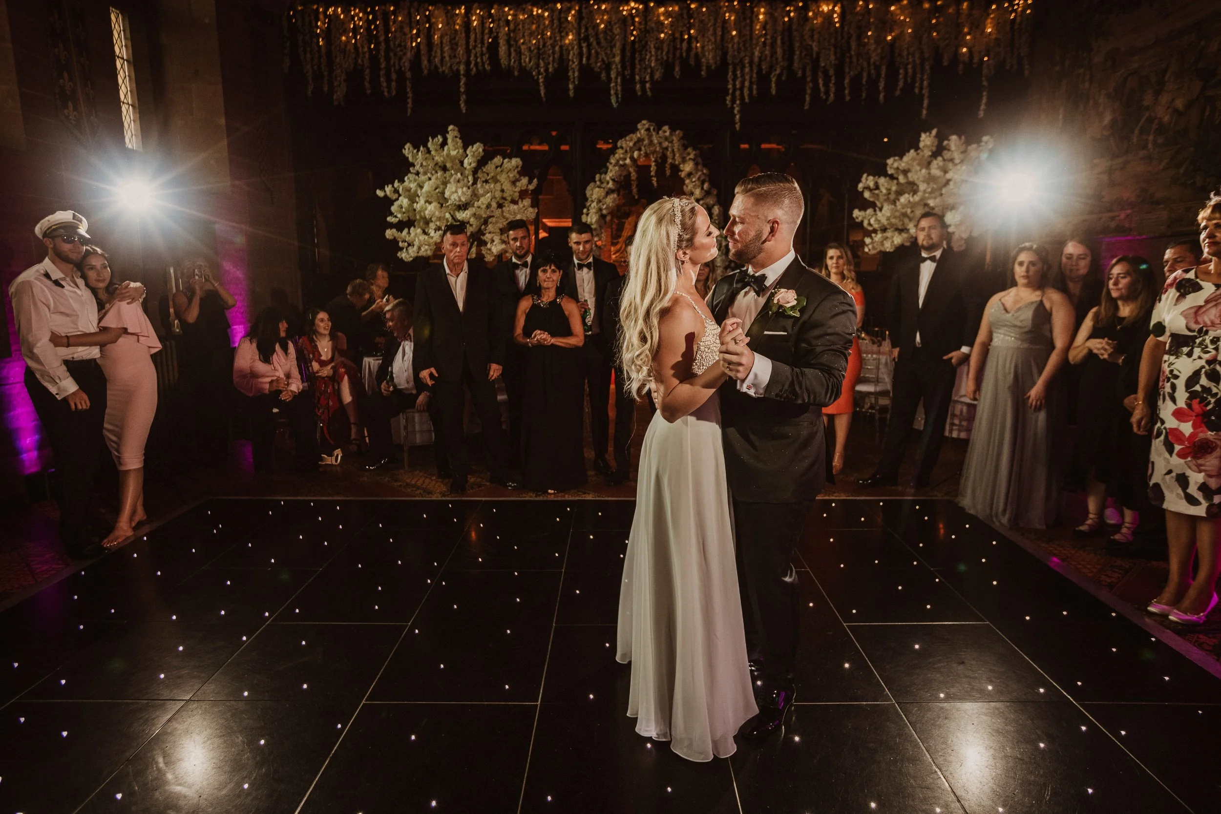 Couple dancing at their wedding reception with guests watching in a dimly lit decorated venue.
