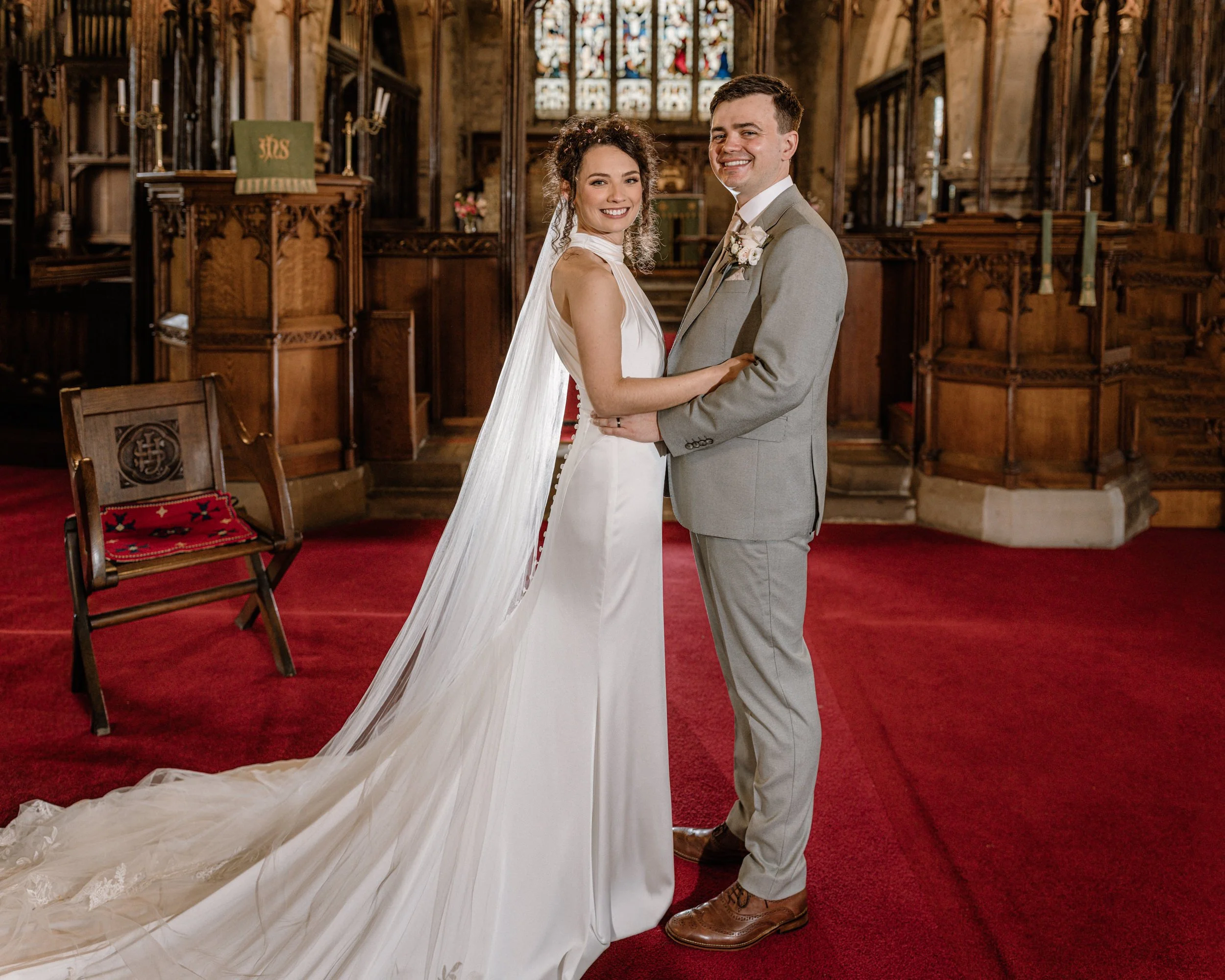 A bride and groom standing inside a church, smiling and holding hands, with the bride wearing a white gown and veil, and the groom wearing a light gray suit. The church features stained glass windows and wooden pews.