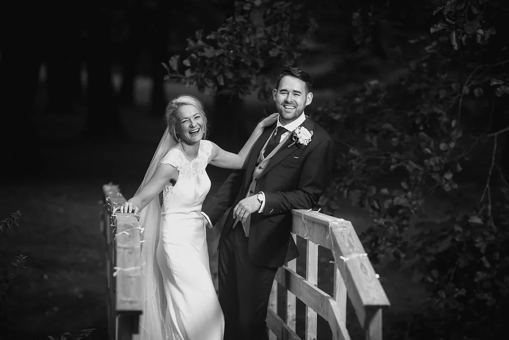 Black and white photo of a happy bride and groom standing on a wooden bridge outdoors, smiling and laughing.