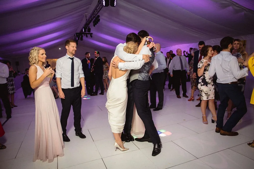 People dancing and hugging at a wedding reception under a purple-lit tent.