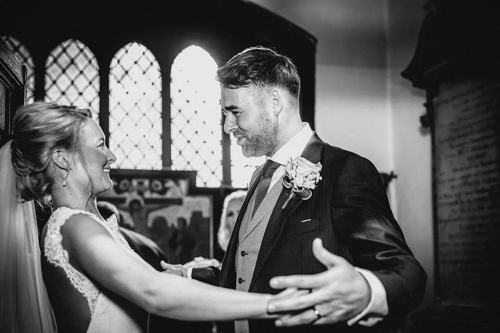 A couple in wedding attire dancing in a church, smiling at each other, with stained glass windows in the background.